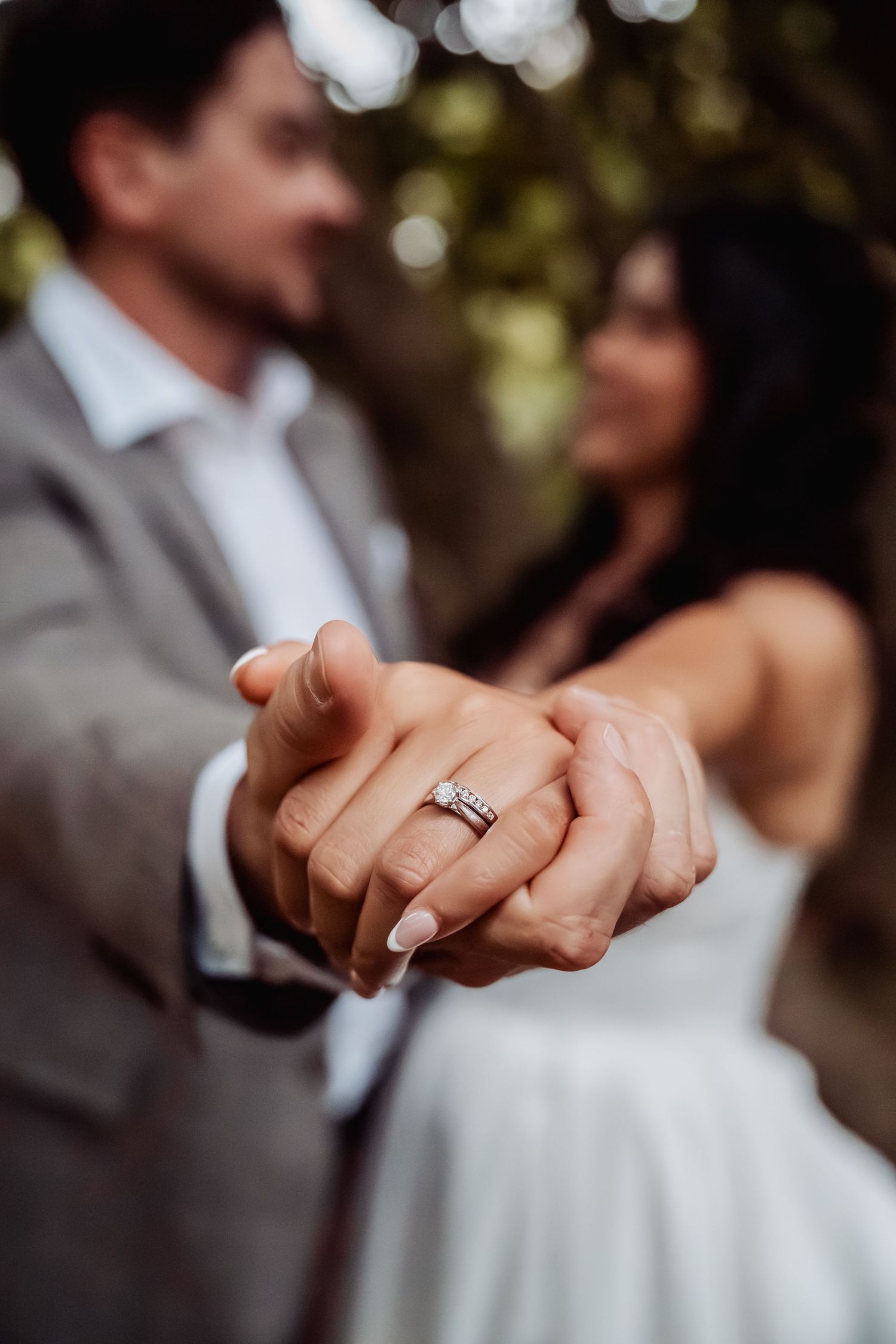 Close-up of bride and groom holding hands showing diamond wedding ring in an outdoor setting.