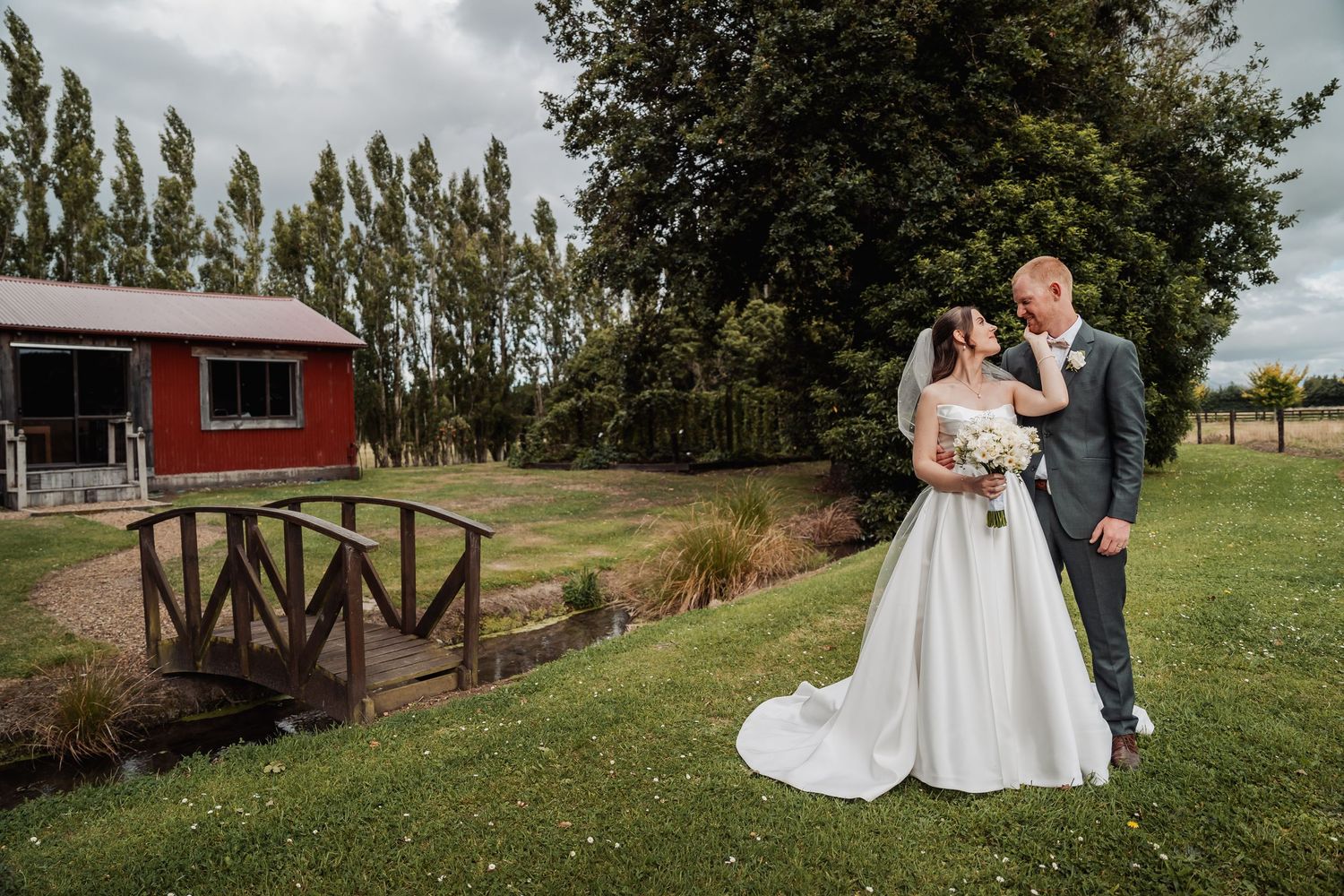 Bride and groom pose on lush green lawn near red barn and wooden bridge at scenic outdoor wedding venue.