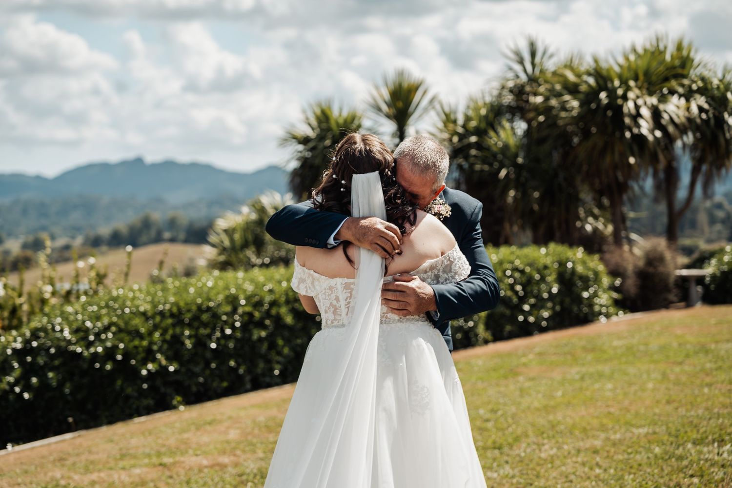 Bride in white gown sharing an emotional hug with older man outdoors with tropical greenery behind them.