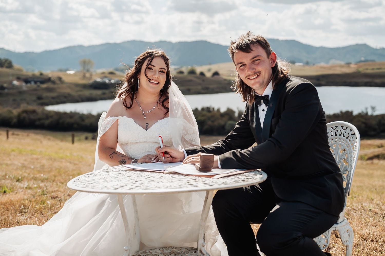 Smiling bride and groom signing wedding documents at ornate white table with lake and mountains behind.