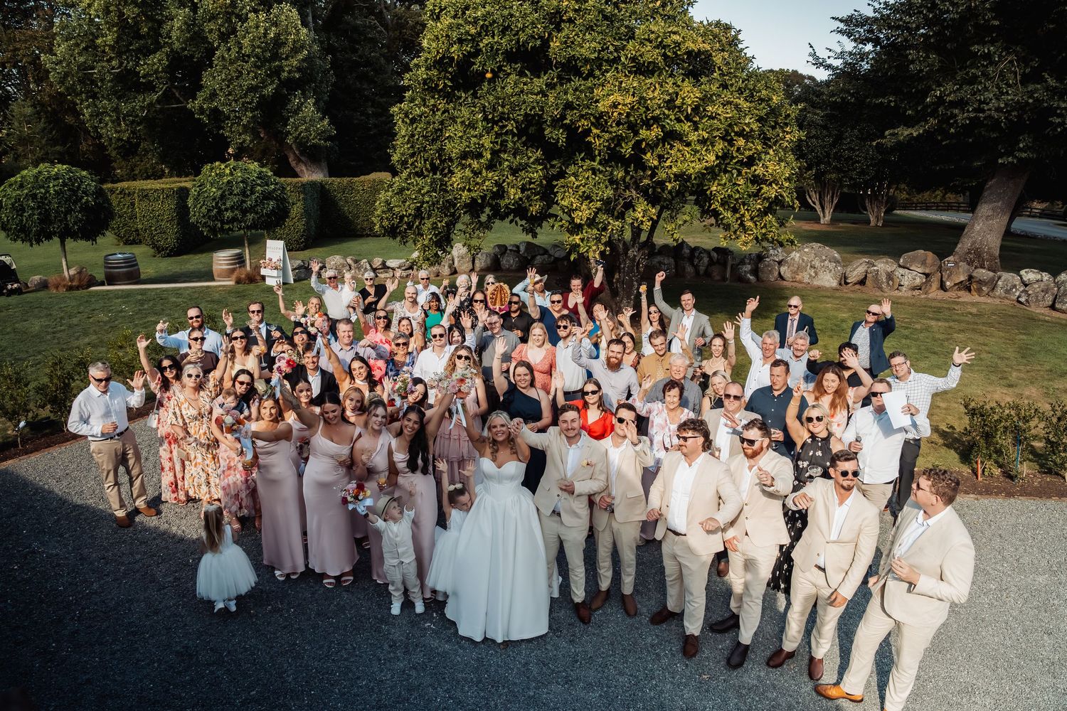 Large wedding group photo with bride, groom, bridesmaids, and guests celebrating outdoors.