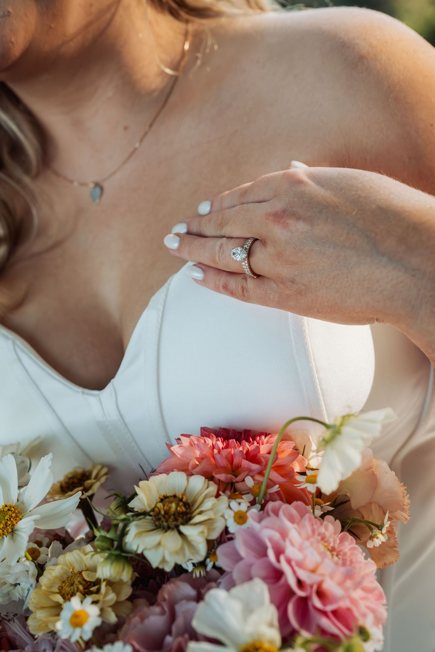 Bride in white dress displaying diamond engagement ring while holding a colorful floral bouquet with pink and white flowers.