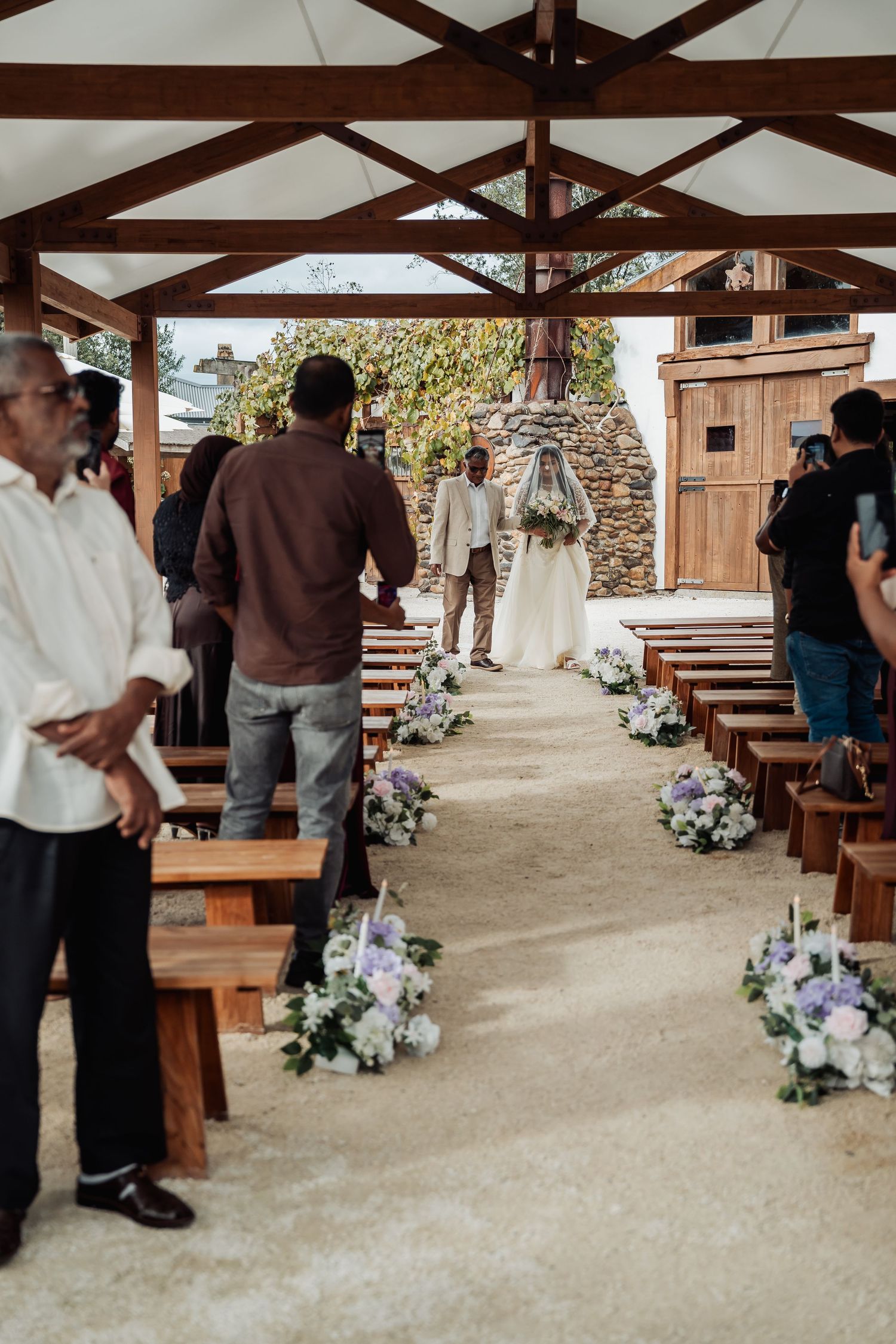 Wedding ceremony aisle lined with floral arrangements under wooden pavilion with bride and groom at altar.