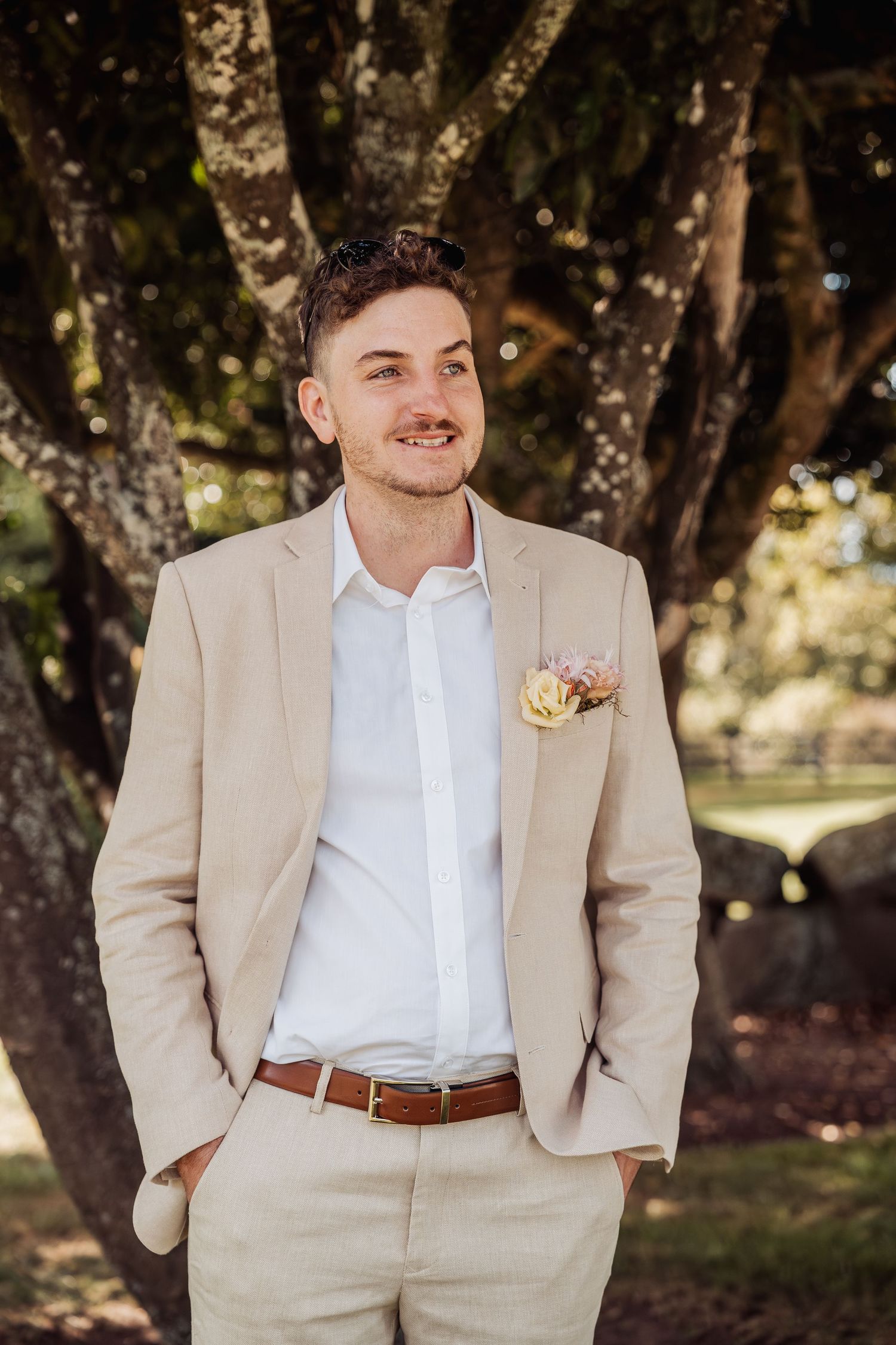 Handsome groom in a beige suit with floral boutonniere standing outdoors under a large tree.