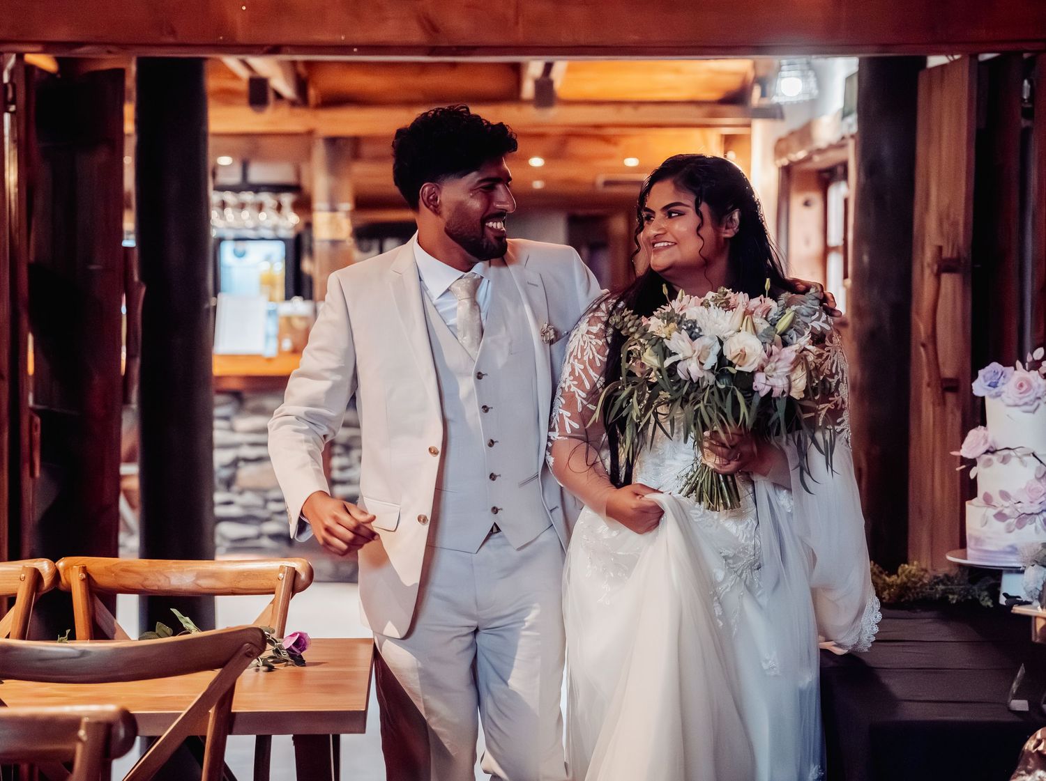 Smiling bride and groom in elegant wedding attire walking together inside rustic wooden venue.