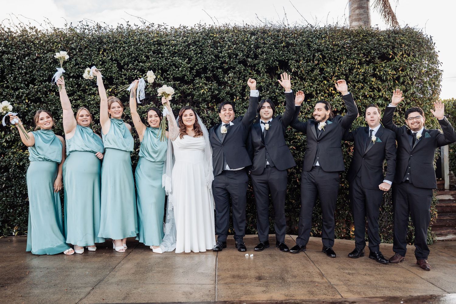 Wedding party poses outdoors with bridesmaids in light blue dresses and groomsmen in dark suits.
