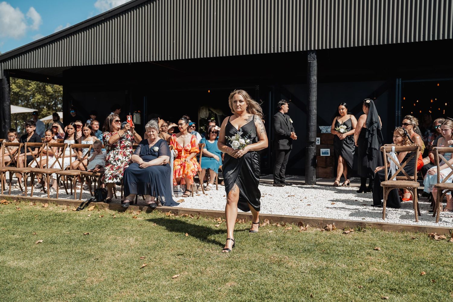 Wedding guest in navy dress walks on grassy lawn past seated audience outside a modern black barn venue.