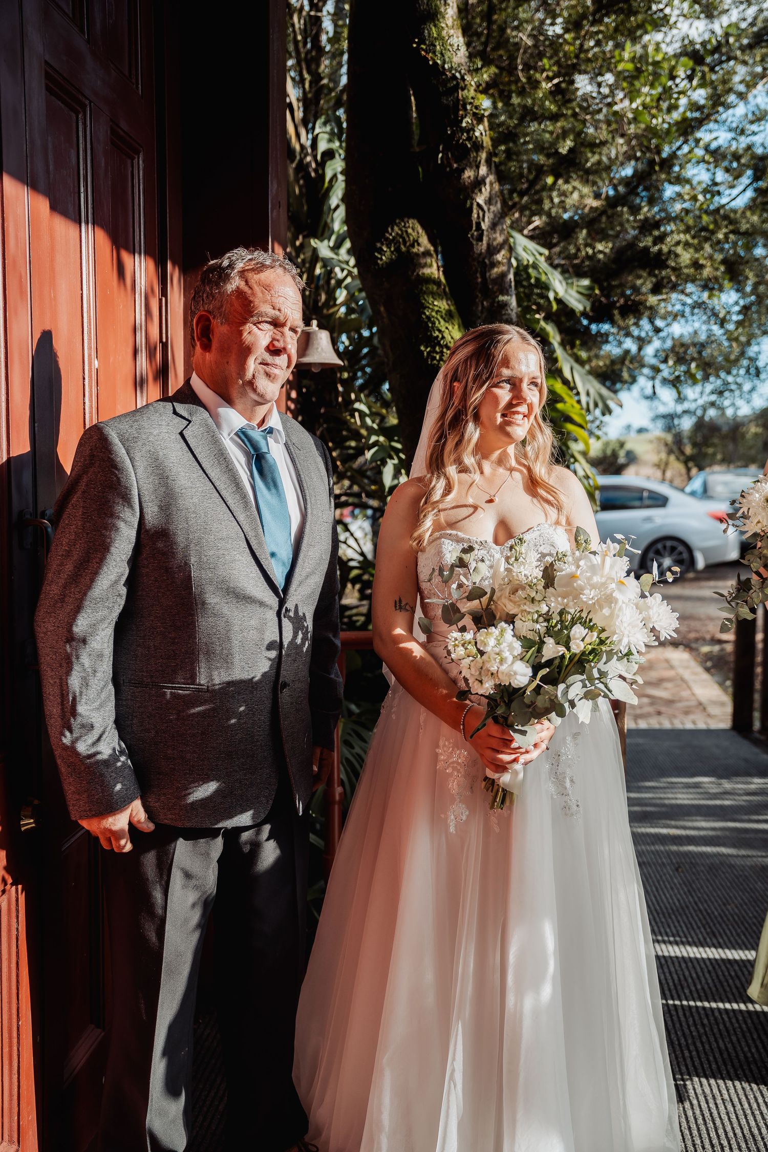 A couple stands together in sunlight outside a red door on their wedding day, with the bride holding a white bouquet.