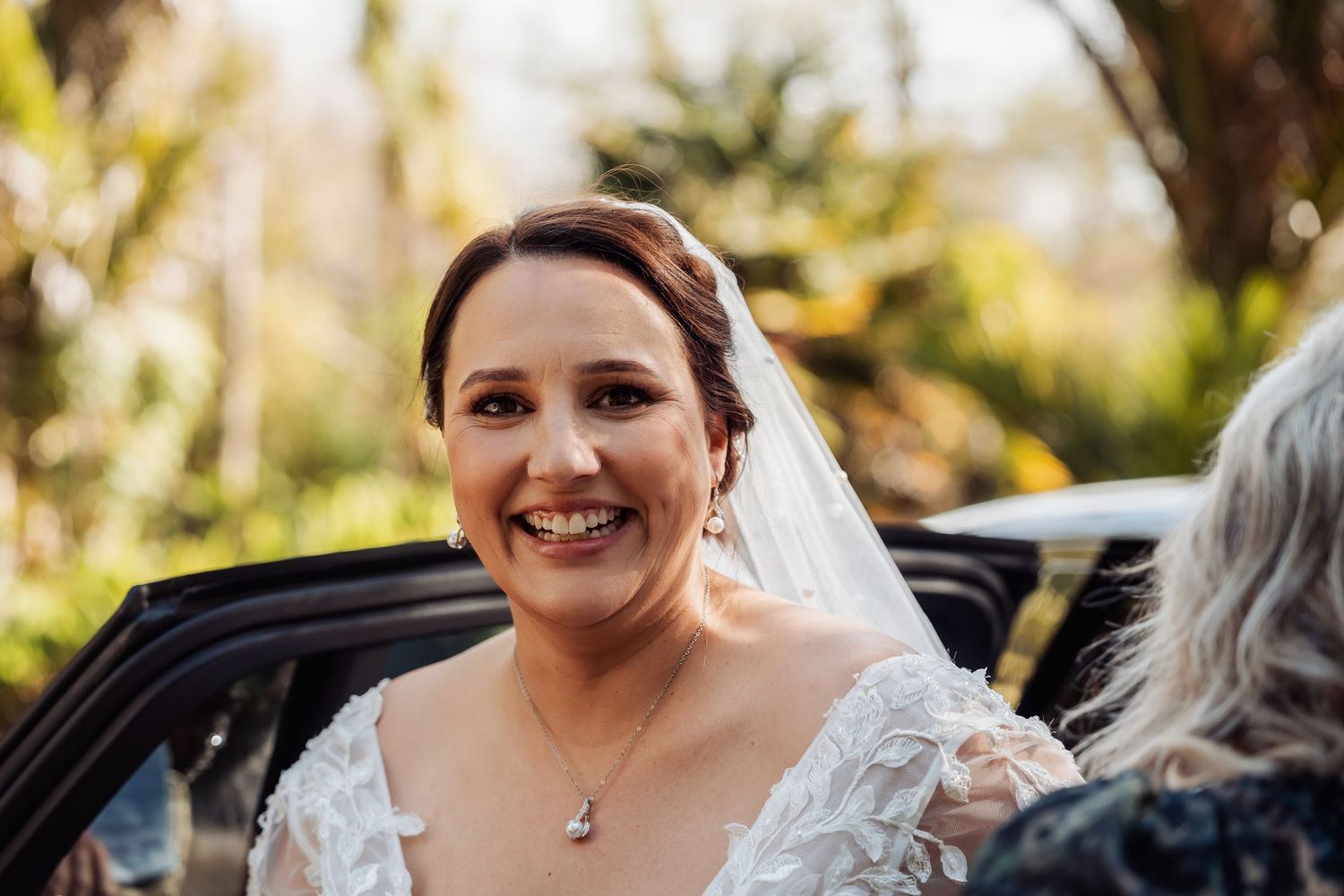 A bride laughs joyfully during an outdoor wedding photoshoot with natural lighting and greenery in the background.