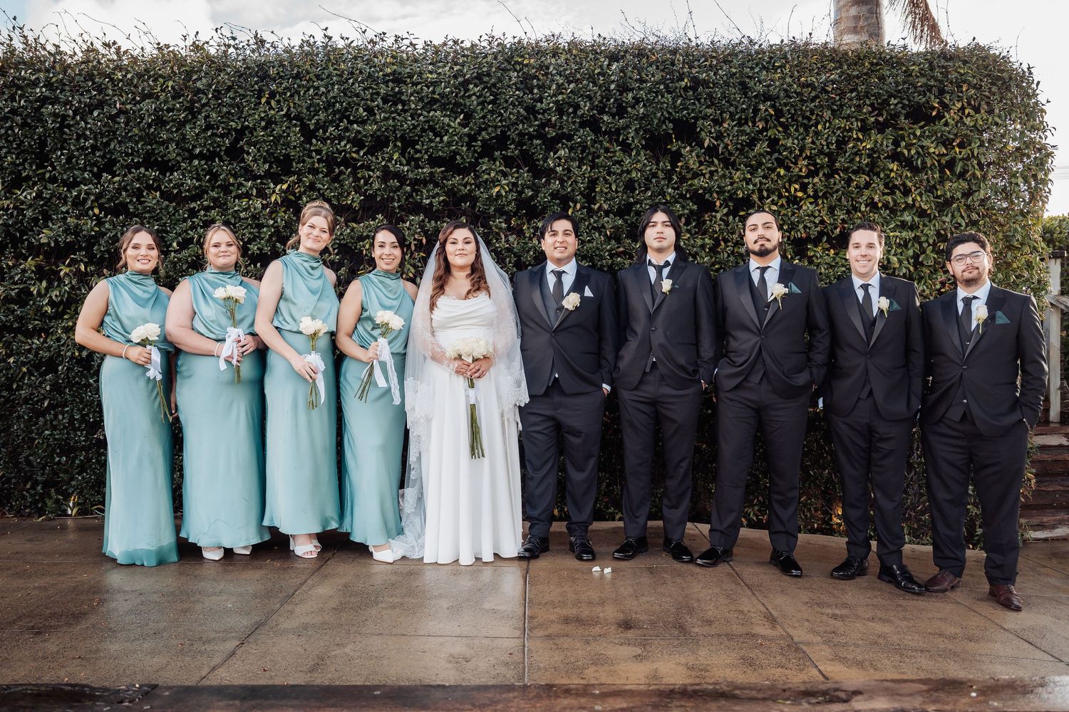 Wedding party poses against a garden wall in light blue bridesmaid dresses and black suits.