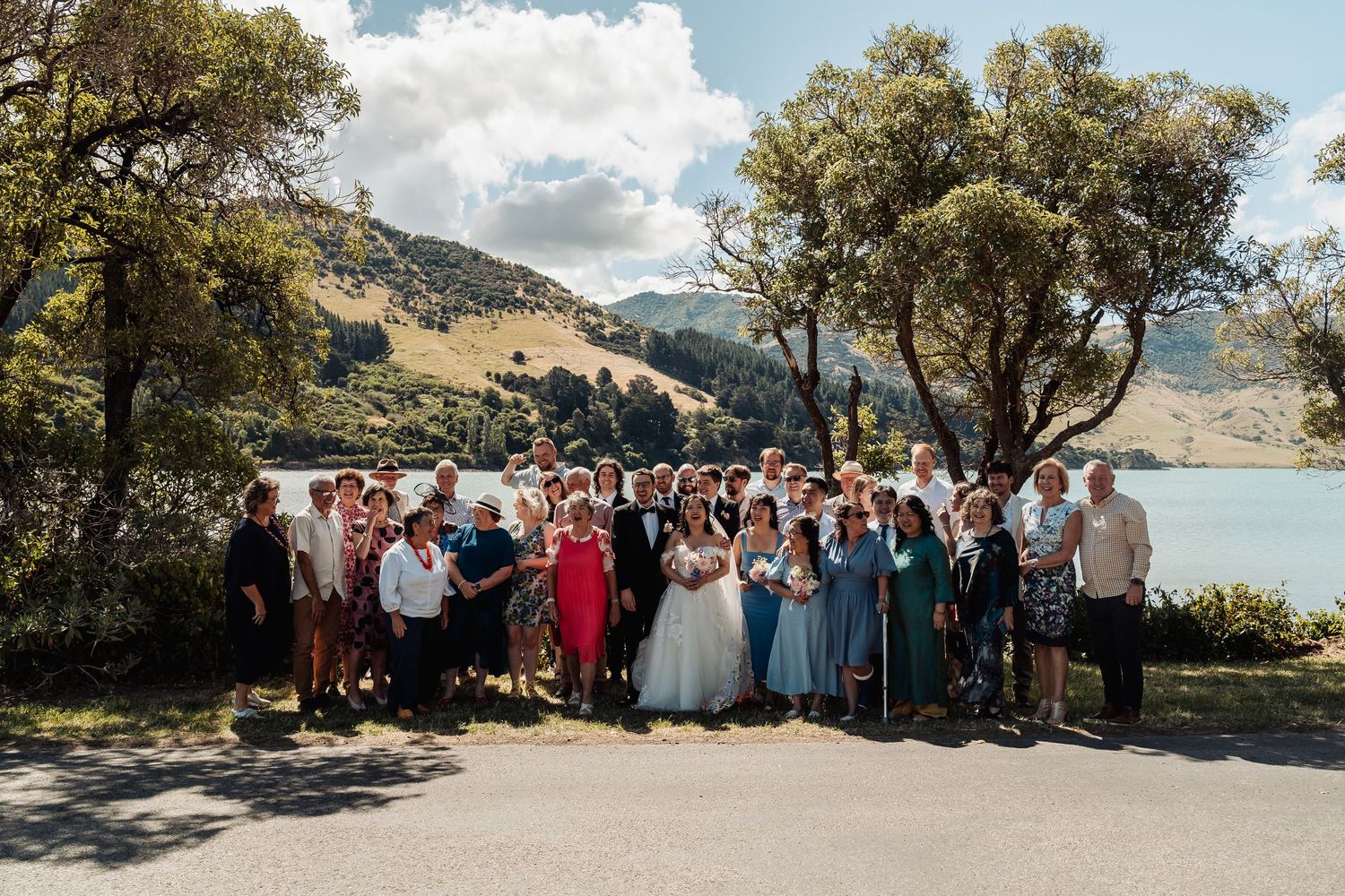 Large wedding party group photo outdoors by a scenic New Zealand lake surrounded by lush green hills.