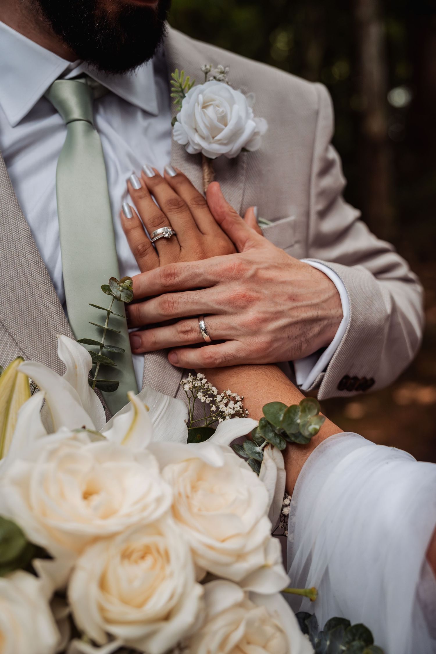 Close-up of bride and groom hands with wedding rings resting on beige suit with white rose boutonniere.