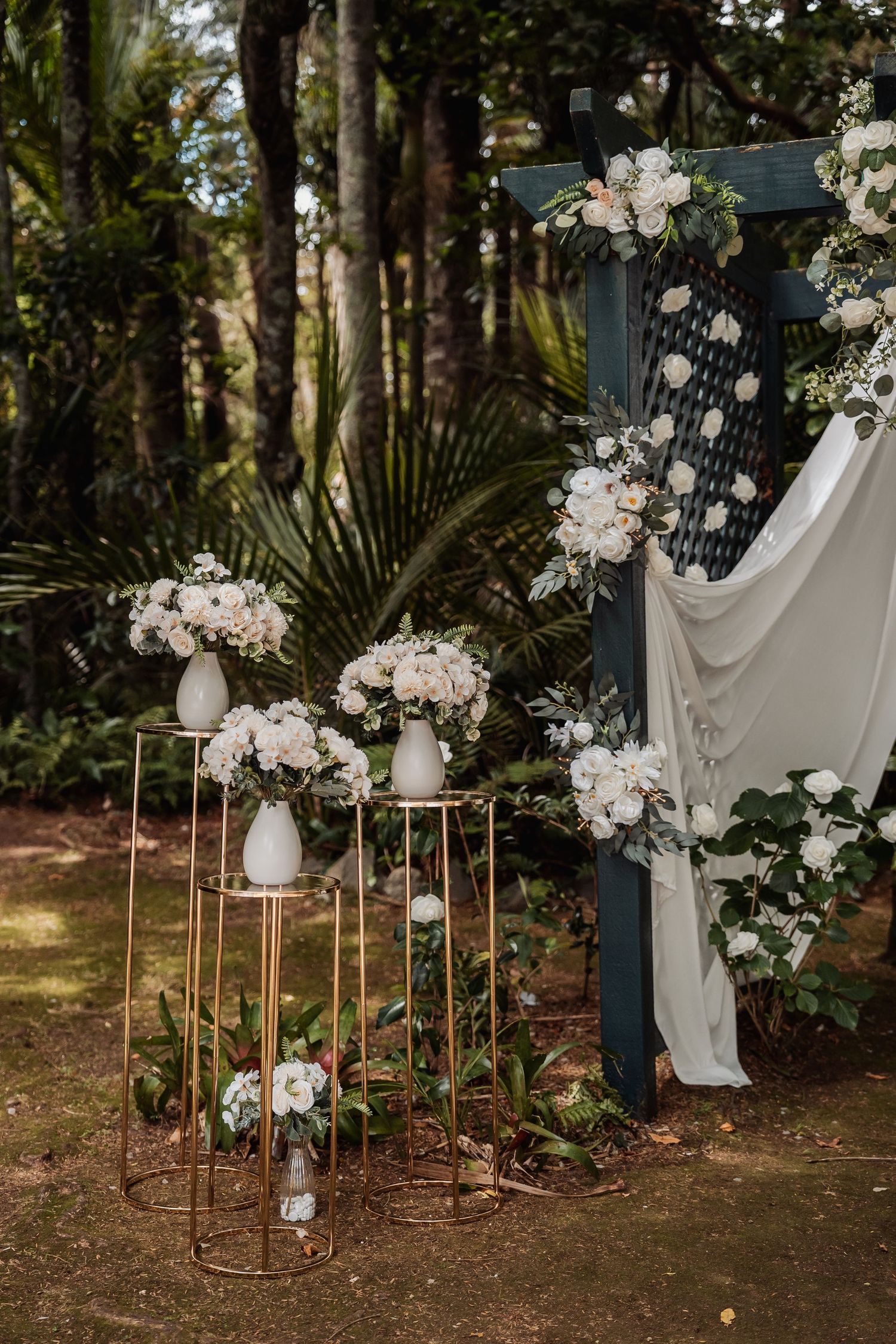 Gold geometric stands with white floral arrangements beside a dark wedding arch draped in white fabric outdoors.