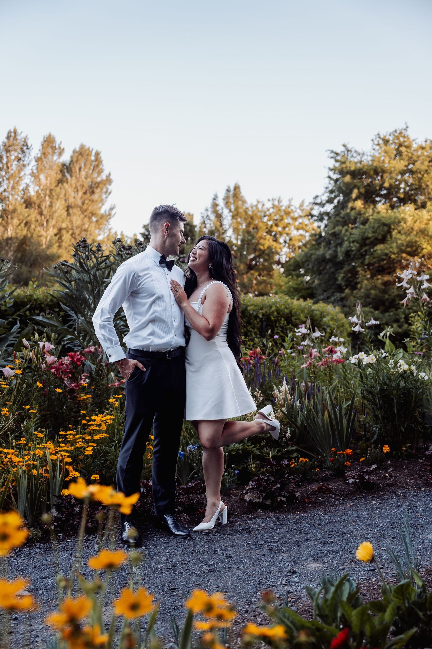 Couple in wedding attire embracing in a colorful garden, bride in white dress and groom in white shirt with bow tie.
