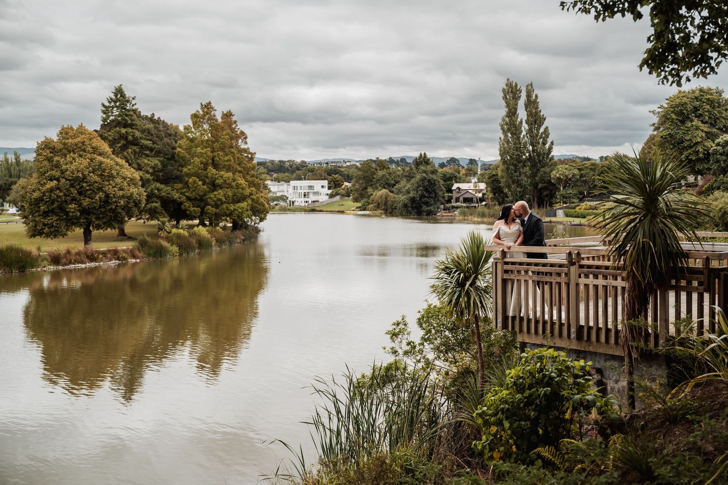 Wedding couple share romantic moment on wooden bridge overlooking serene lake surrounded by lush trees.