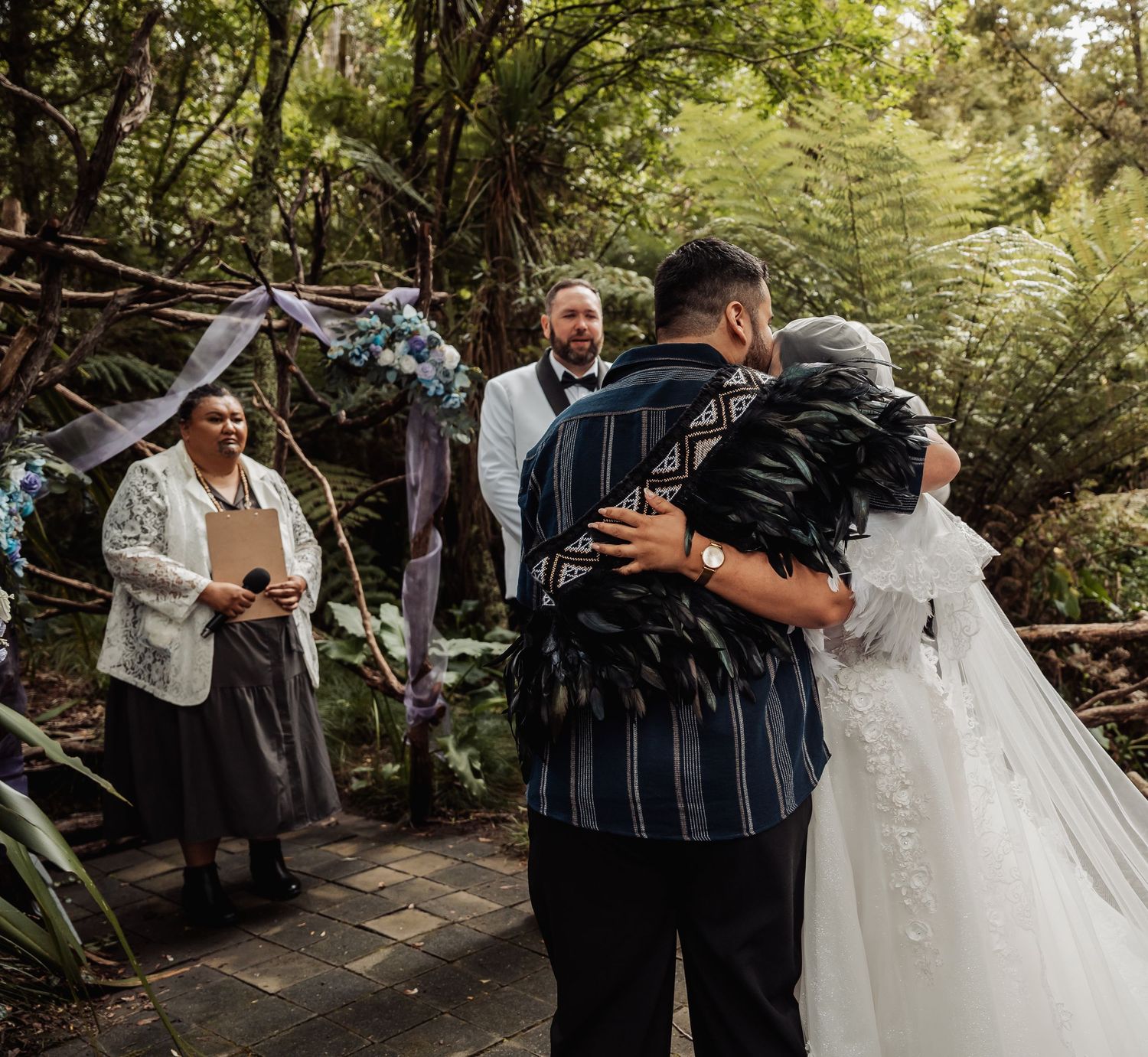 Couple embracing during outdoor wedding ceremony in lush forest setting with floral arch and officiant.