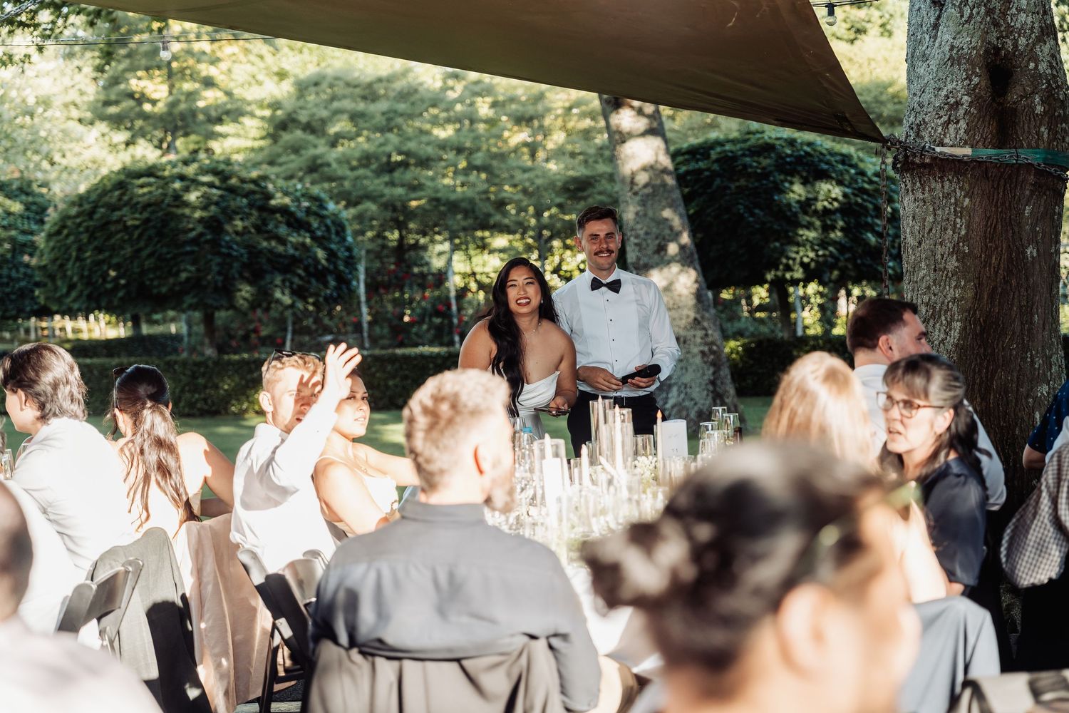 Couple sharing a toast with guests at an elegant outdoor wedding reception surrounded by lush greenery.