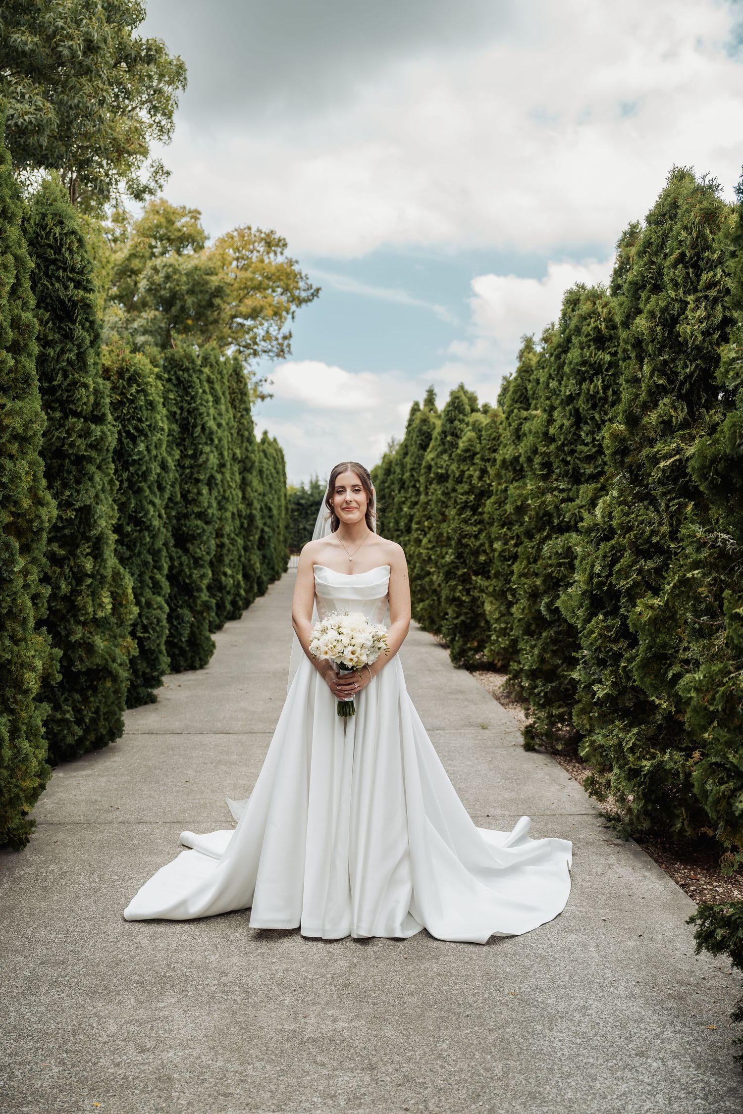 Bride in a white strapless ballgown holding a bouquet stands on a garden path lined with tall evergreen hedges.