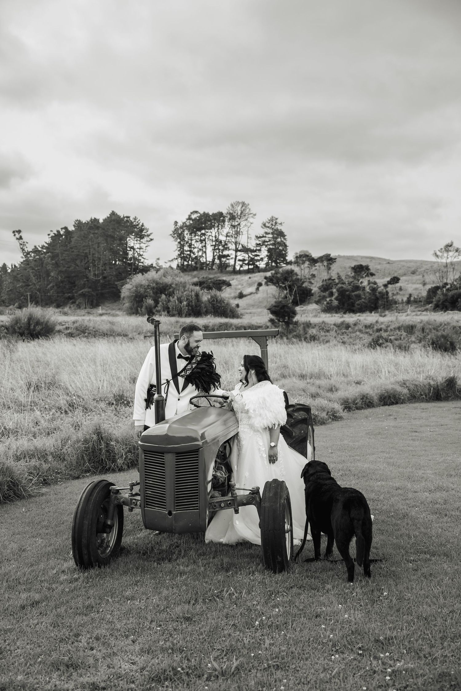 Black and white photo of bride and groom with a black dog beside a vintage tractor in a scenic rural New Zealand landscape.