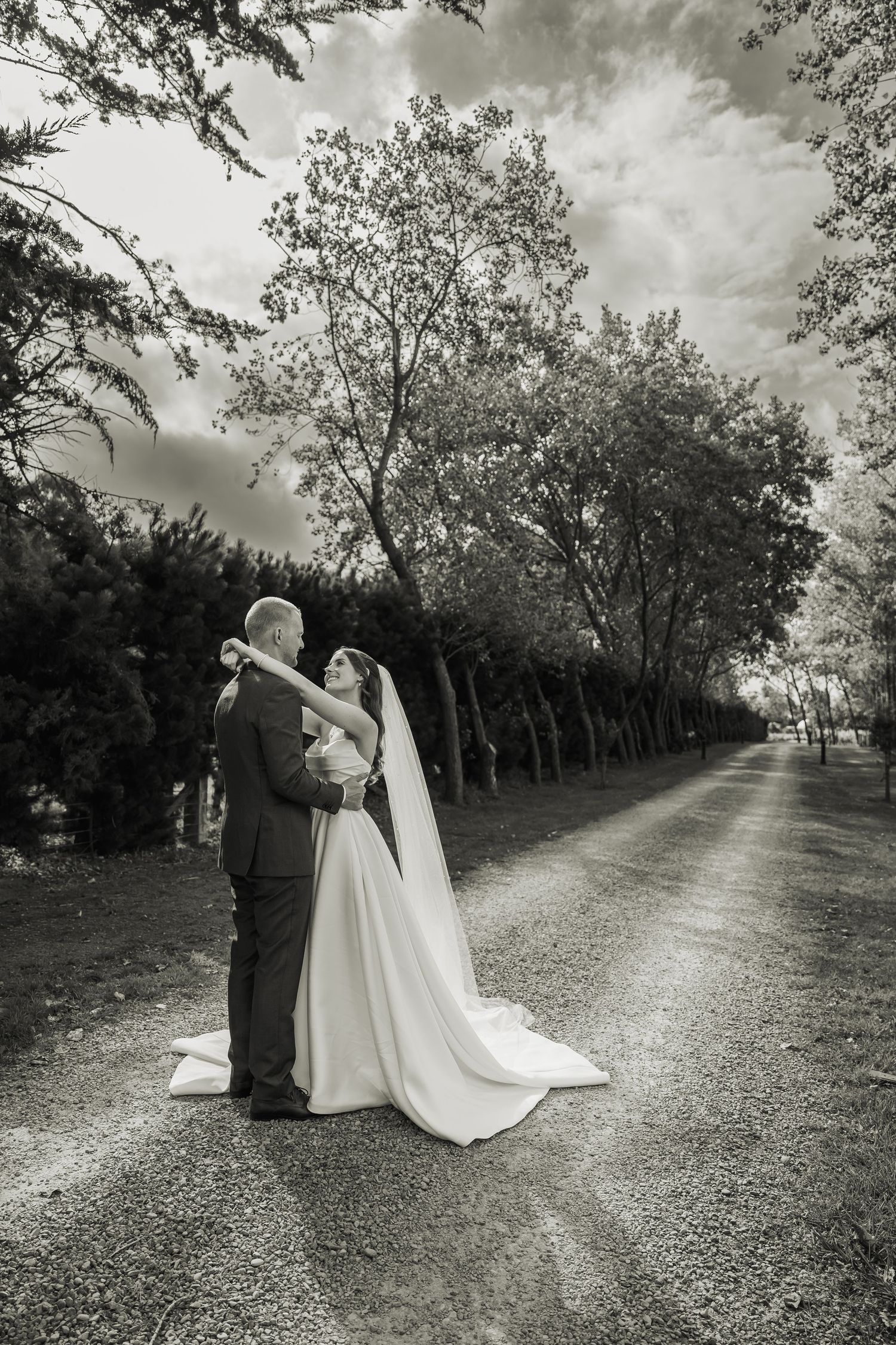Black and white photo of bride and groom embracing on a tree-lined gravel path on their wedding day.