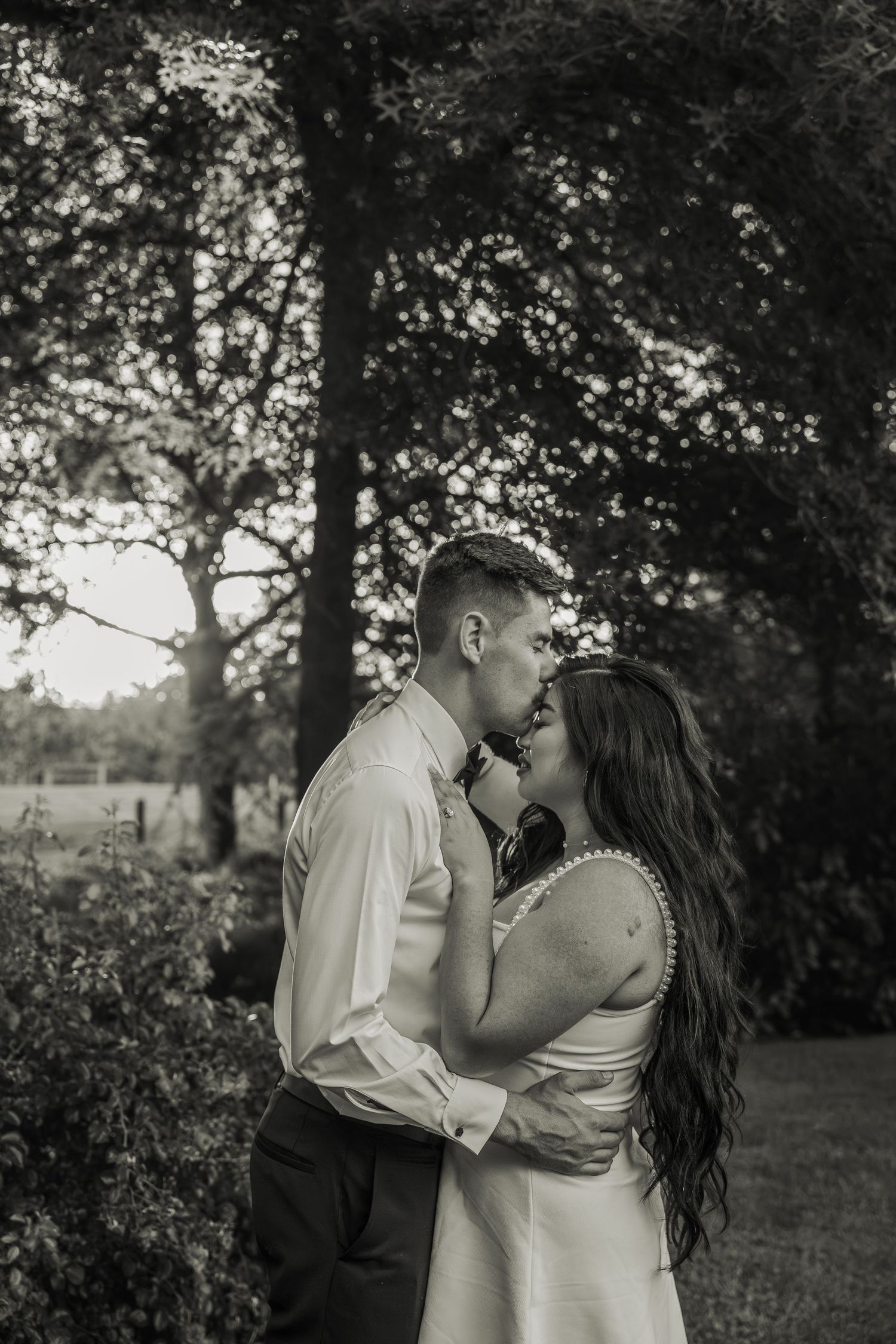 Black and white romantic photo of bride and groom sharing a kiss in lush garden setting.