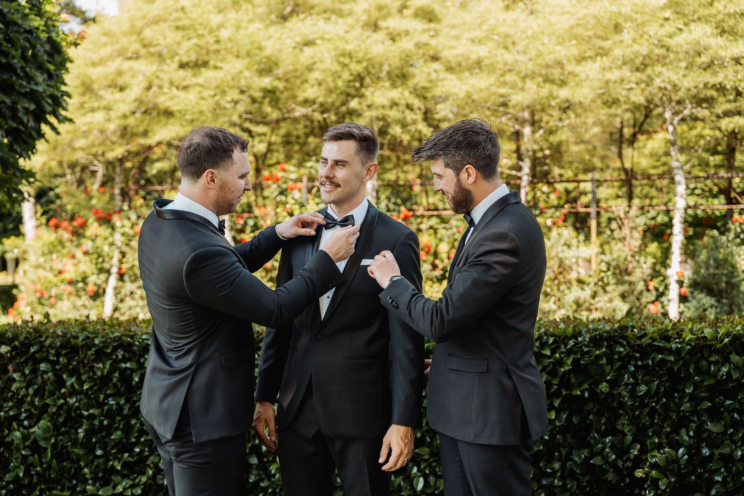 Three groomsmen in dark suits adjust bow ties outdoors in a garden setting before a wedding ceremony.