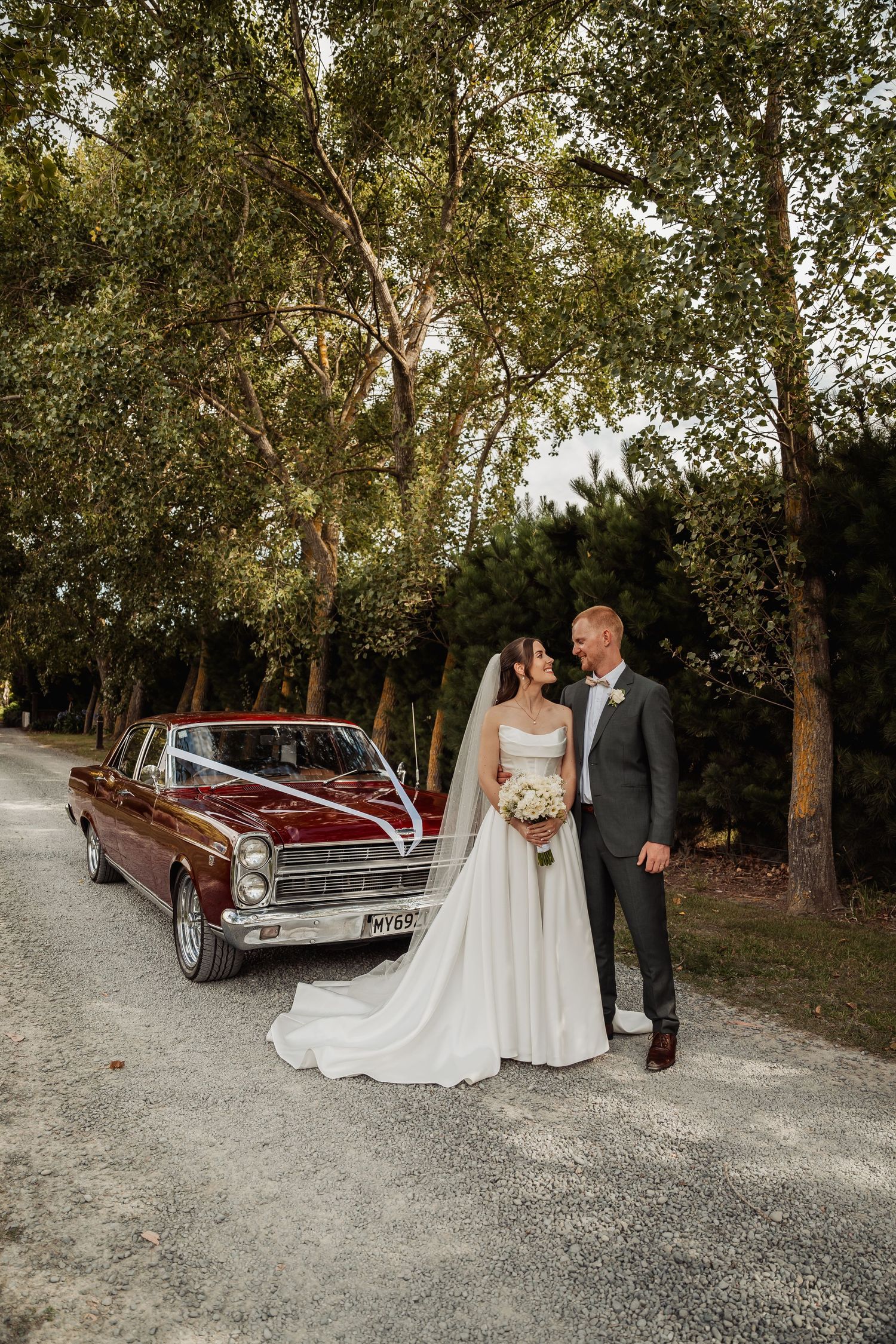 Bride and groom share a tender moment beside a classic red vintage car on a tree-lined gravel driveway.