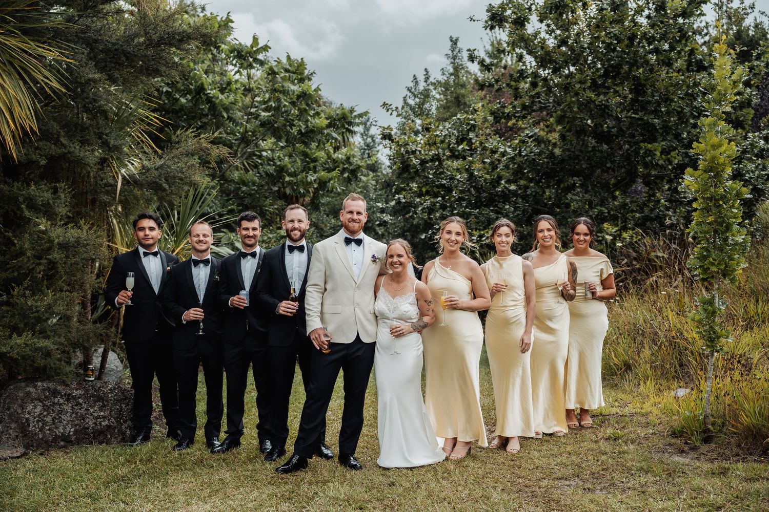 Wedding party posing outdoors with groom in white jacket, groomsmen and bridesmaids in gold.