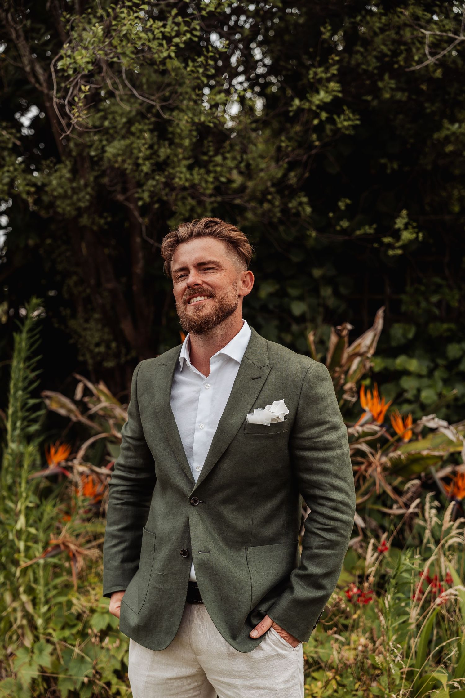 Smiling groom in green blazer and white shirt standing outdoors among tropical garden foliage.