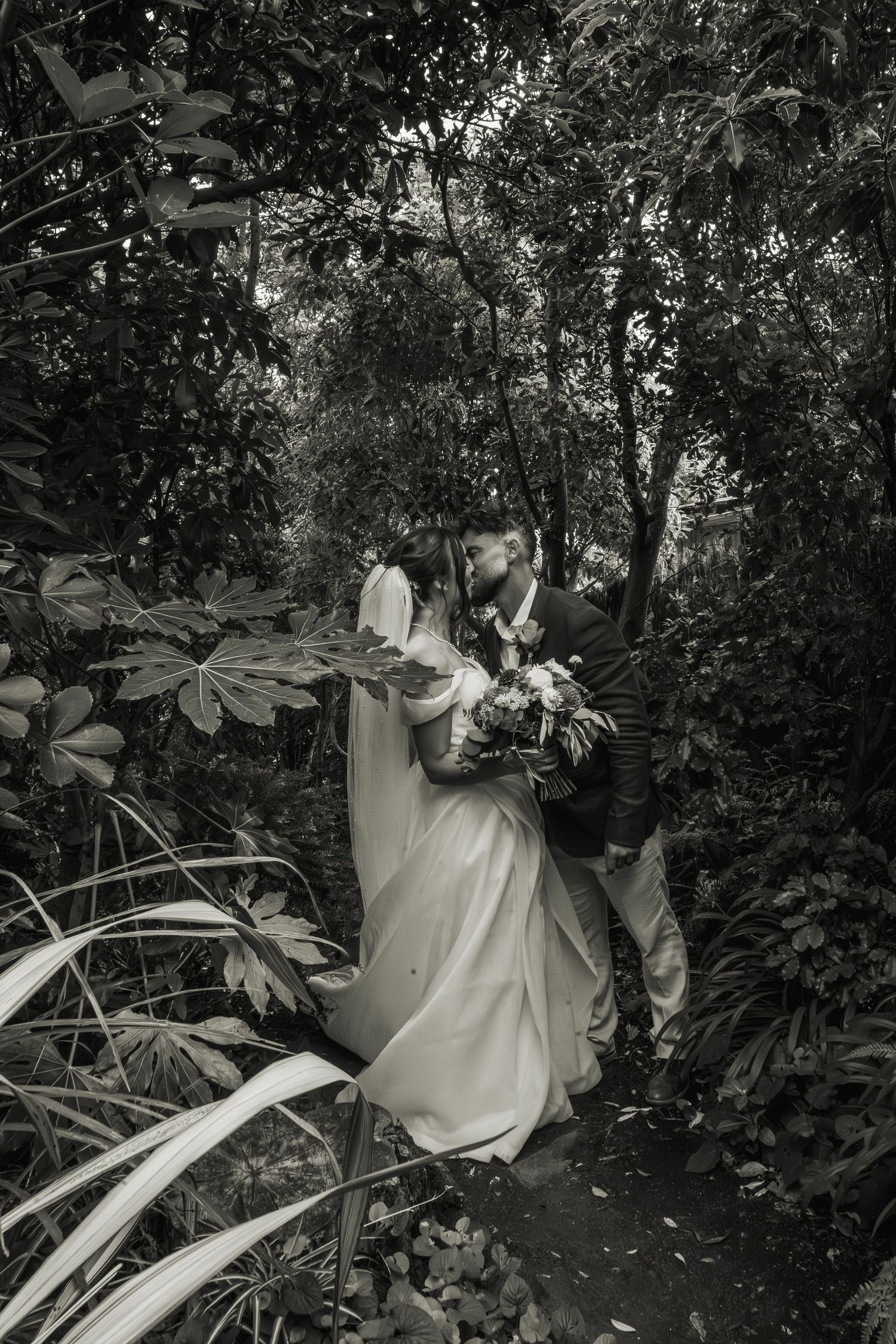 Black and white photo of bride and groom kissing romantically among lush tropical garden foliage.