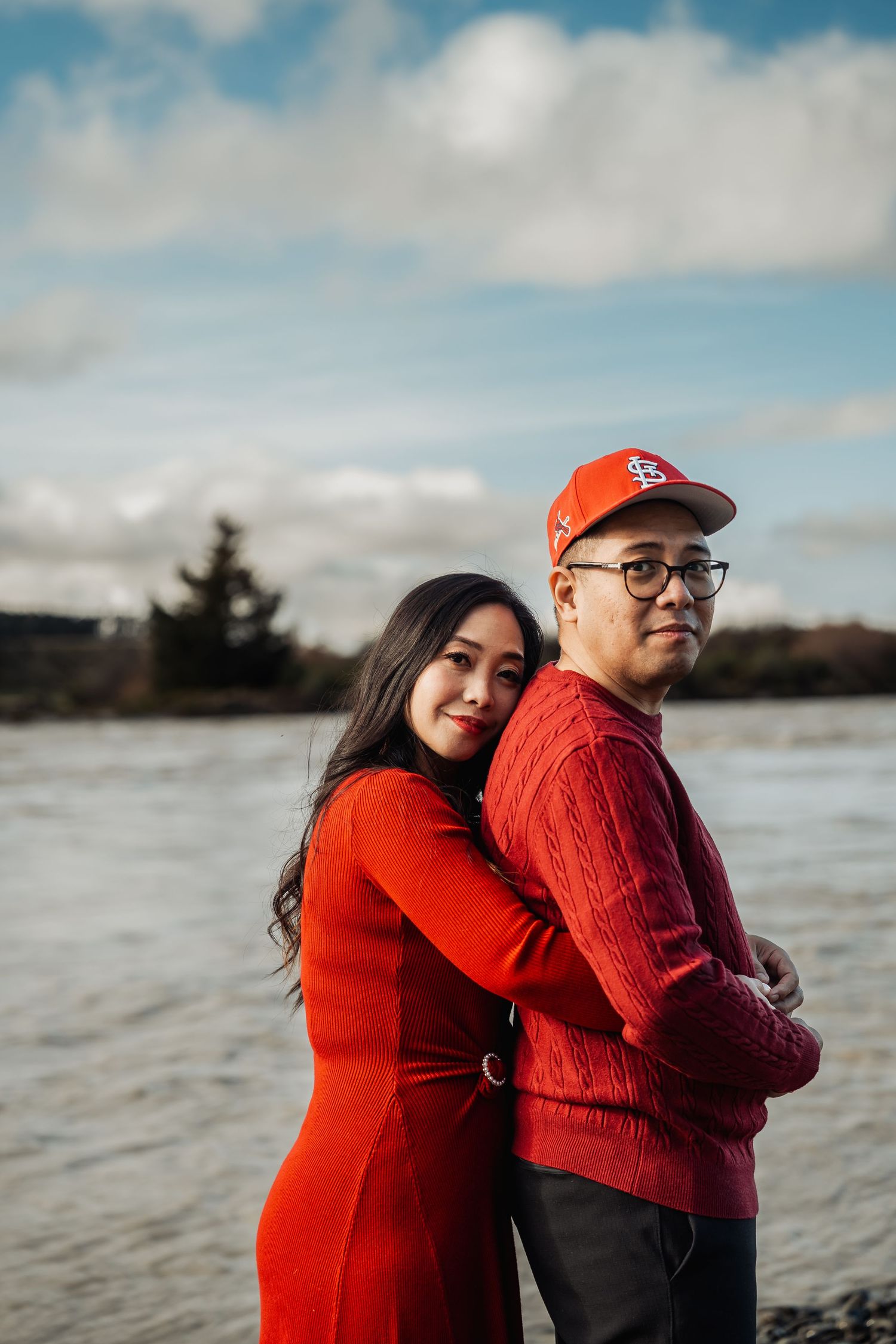 Woman in red dress hugs man in red sweater and cap from behind near a scenic waterfront.