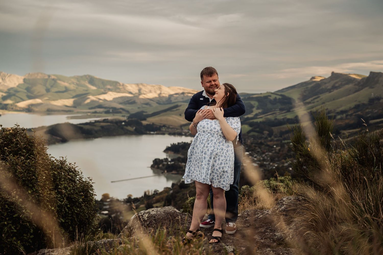 Couple embracing on a rocky hilltop with stunning coastal and mountain views in New Zealand.
