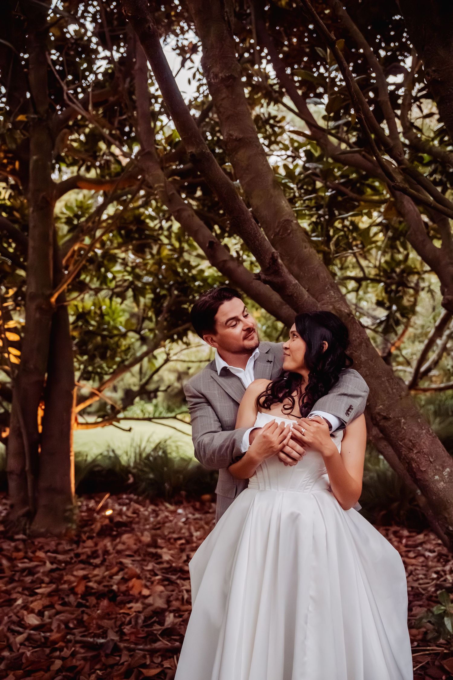 Groom in gray suit embraces bride in white gown under trees with golden autumn leaves in outdoor setting.