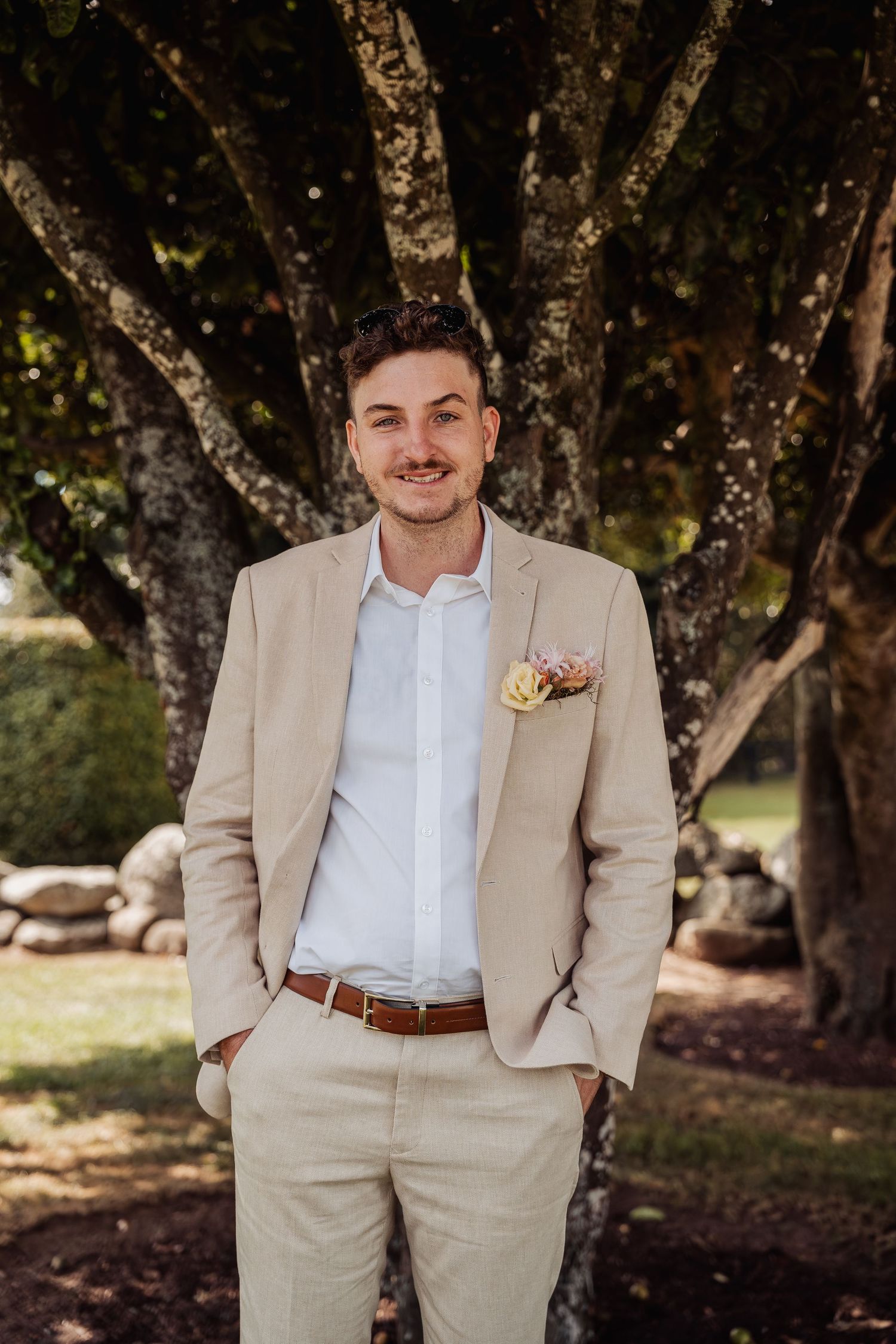 Handsome groom in a beige suit with floral boutonniere smiling outdoors near large trees.