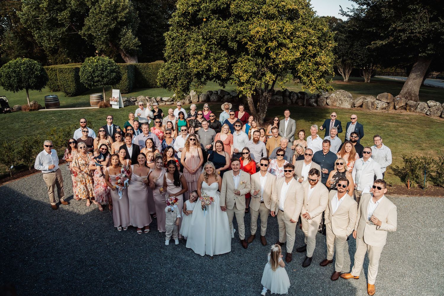 Large wedding group photo with bride, groom, and guests gathered outdoors on sunny day.