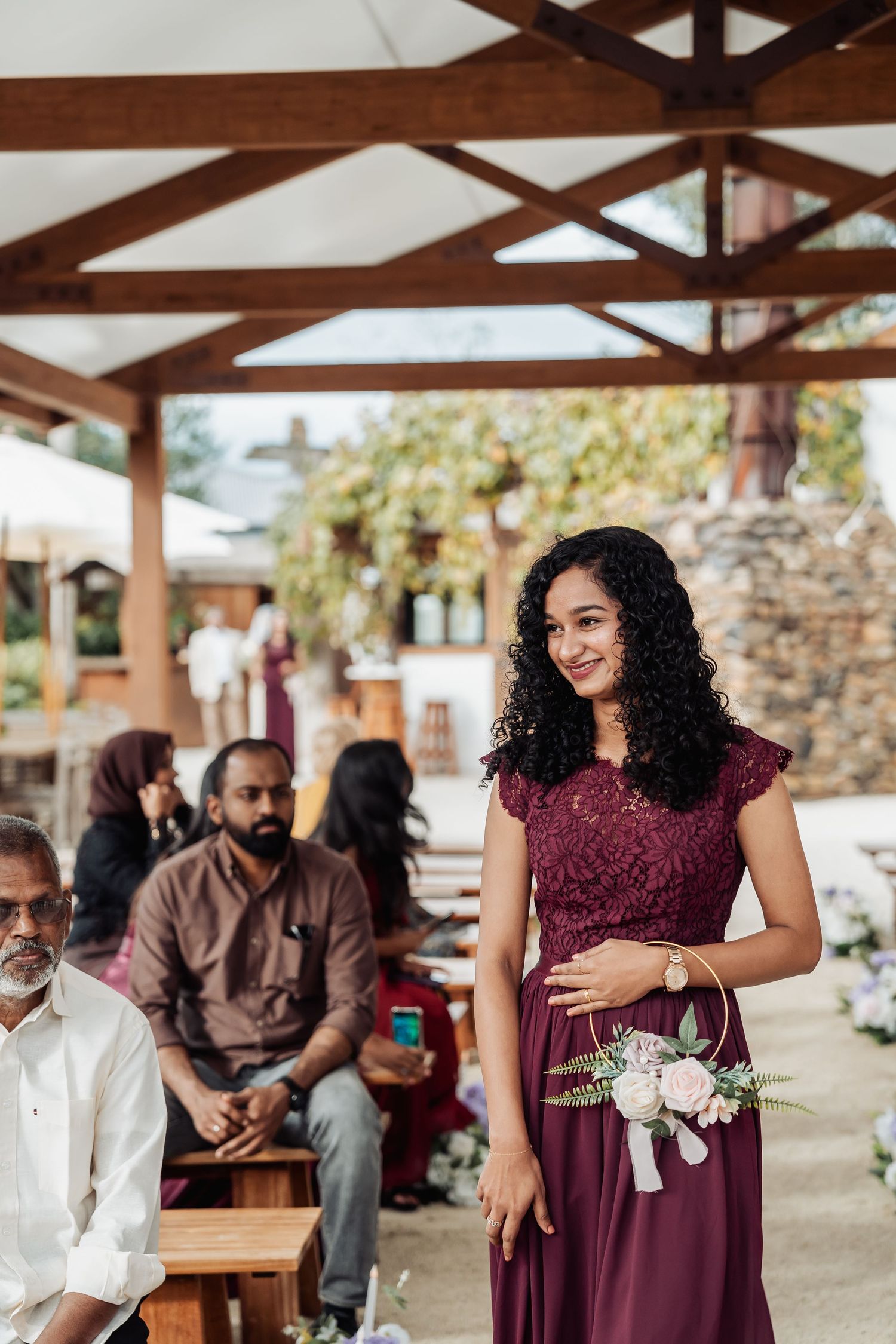 Bridesmaid in burgundy lace dress holding floral hoop bouquet at wedding ceremony.
