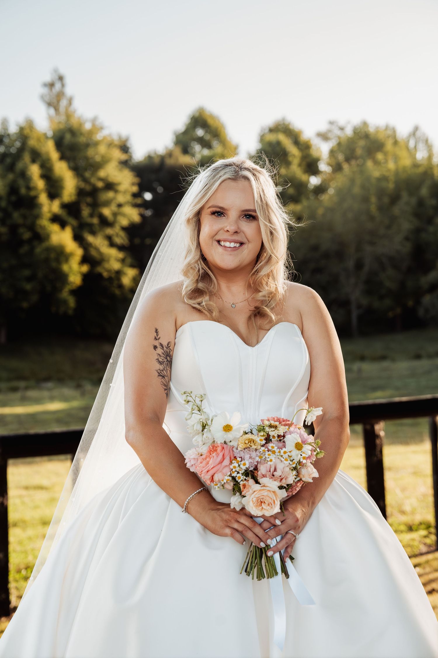Smiling bride in strapless white gown holds a pastel bouquet outdoors on her wedding day.