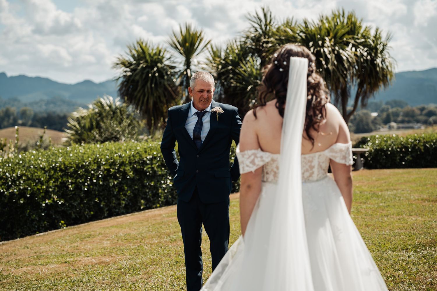Father sees bride in wedding dress for first look moment on green lawn with palm trees and mountains.