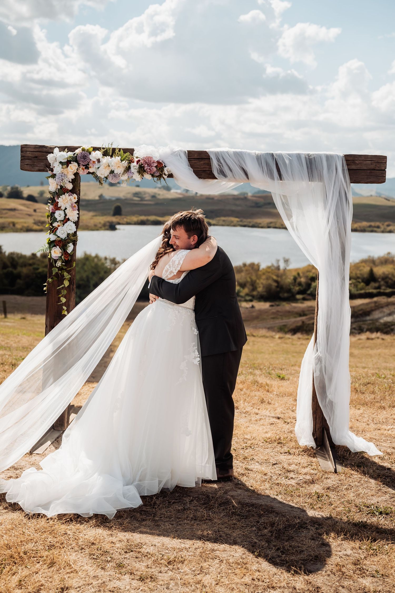 Bride and groom share a romantic embrace under a rustic wooden arch adorned with flowers beside a scenic lake.
