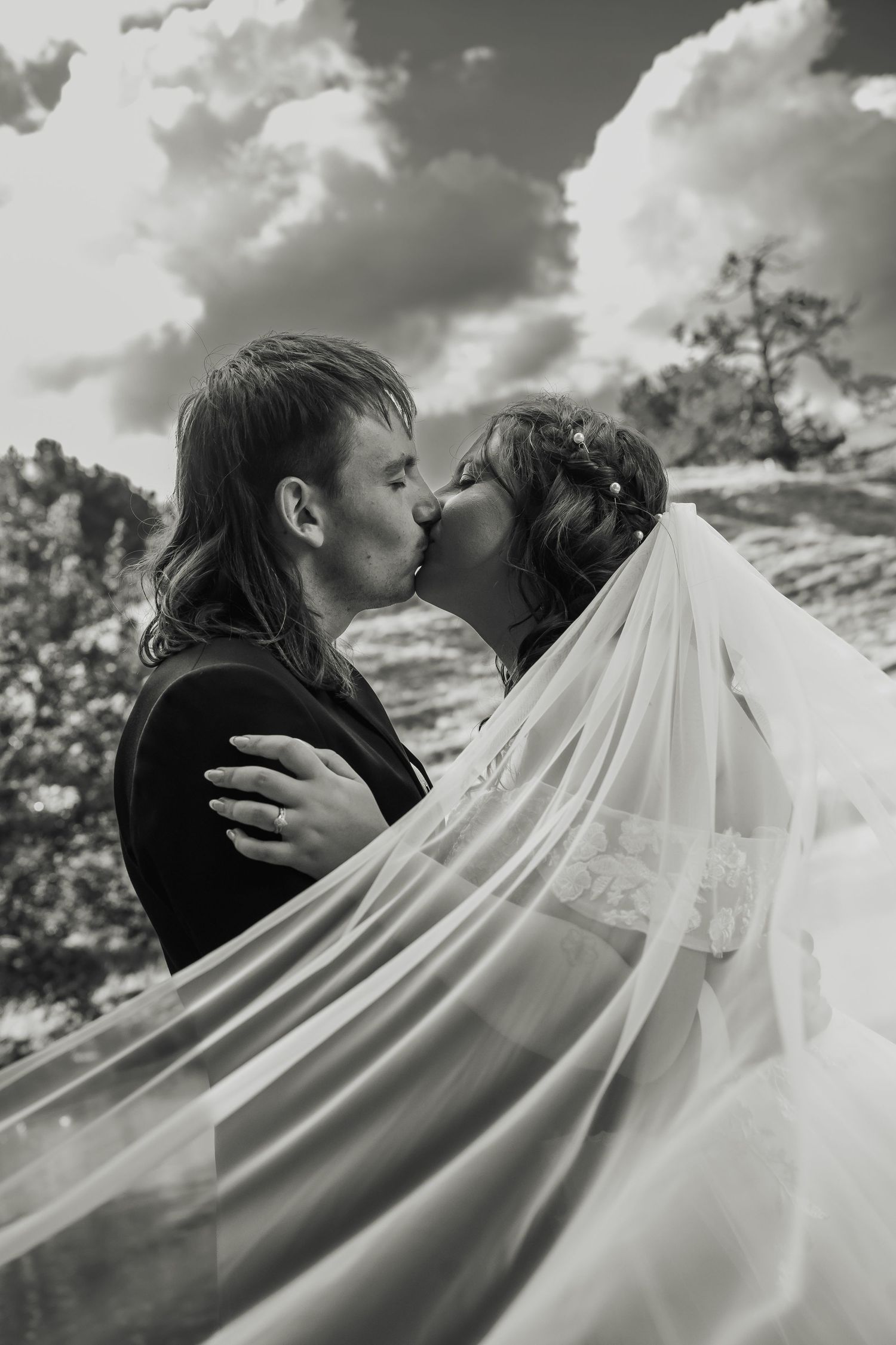 Black and white photo of bride and groom kissing under a flowing veil against a cloudy sky.