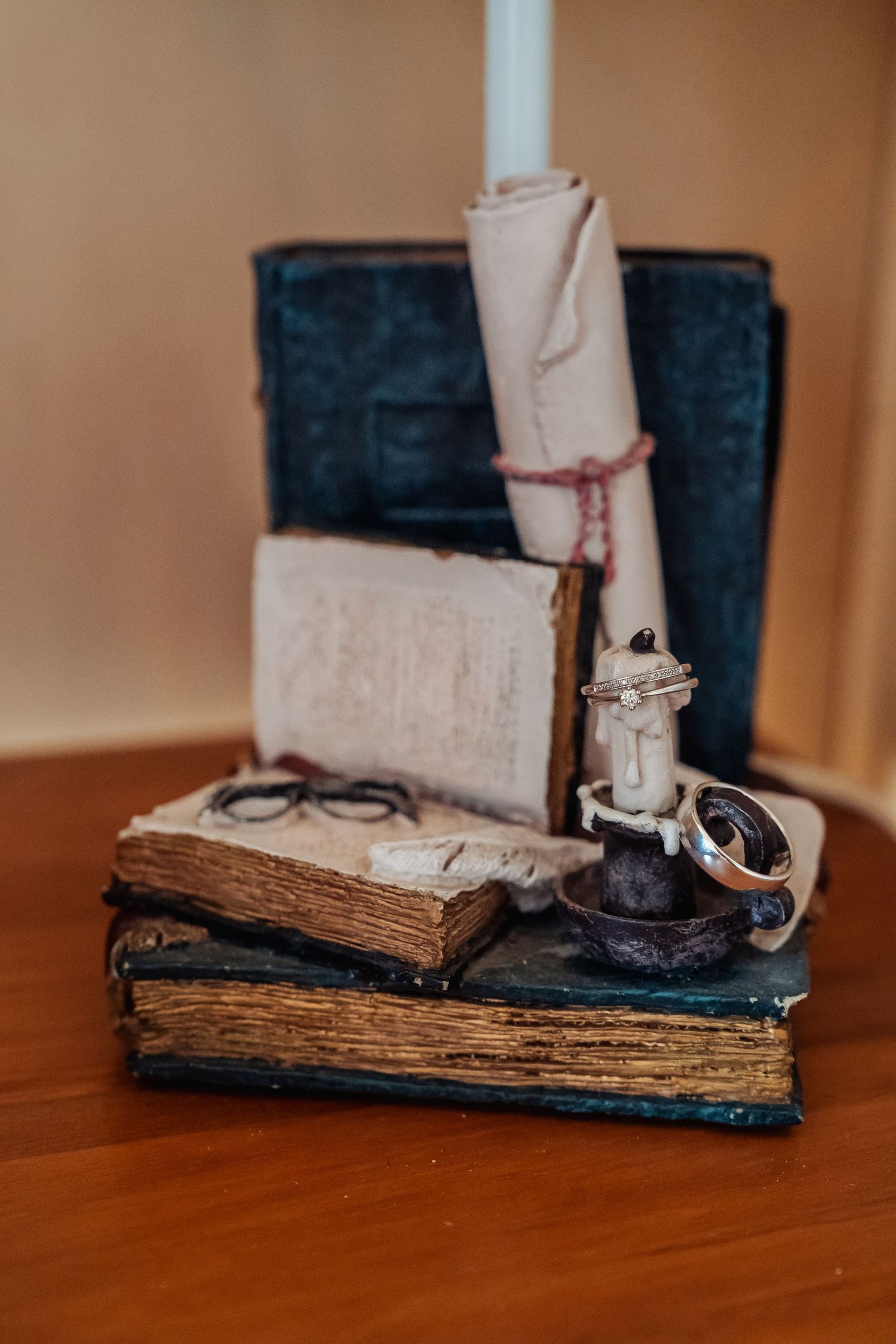 Antique books, rolled parchment scroll, vintage glasses, and wedding rings arranged on a wooden table.