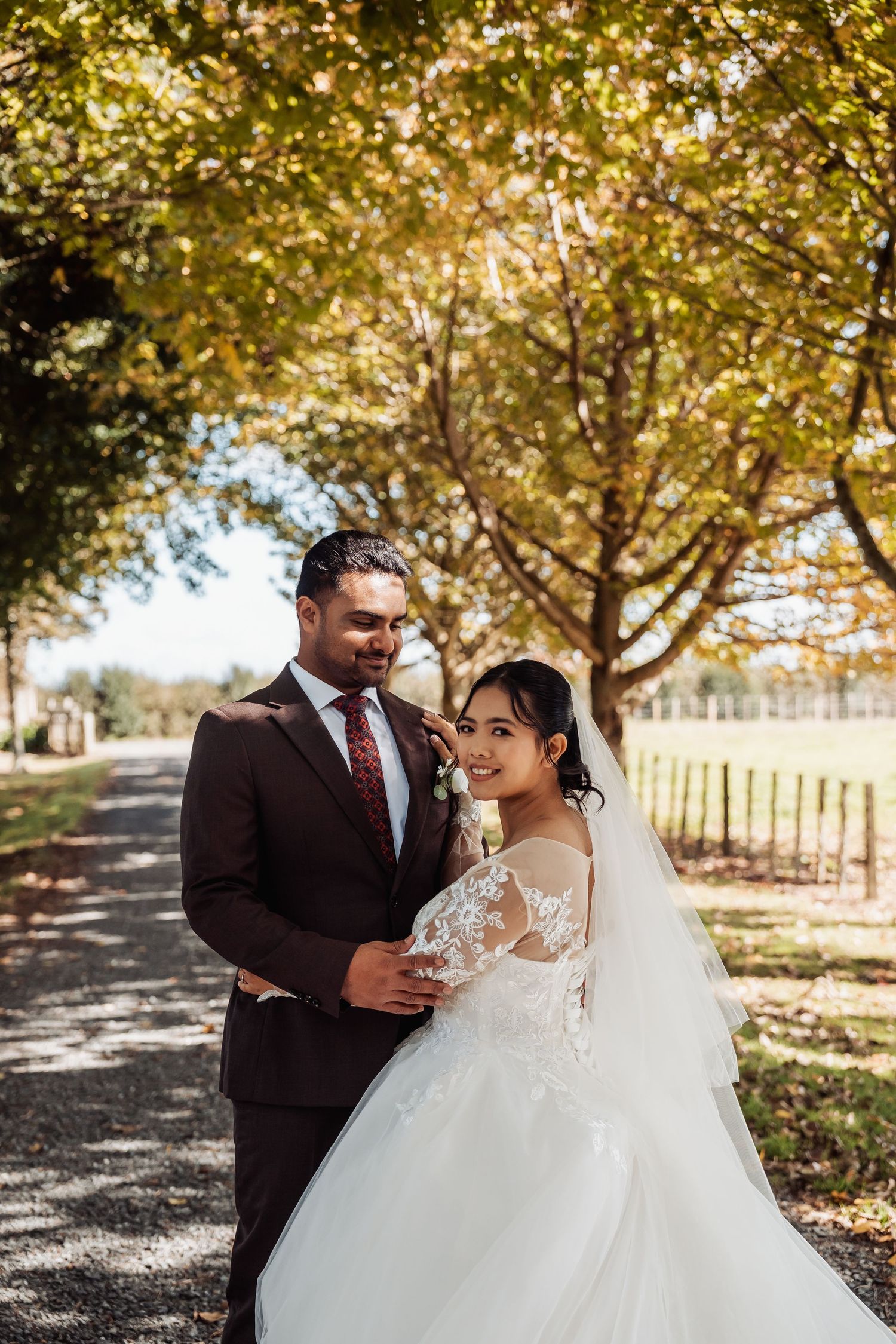 Bride in lace gown and groom in brown suit embrace on tree-lined path in autumn wedding portrait.