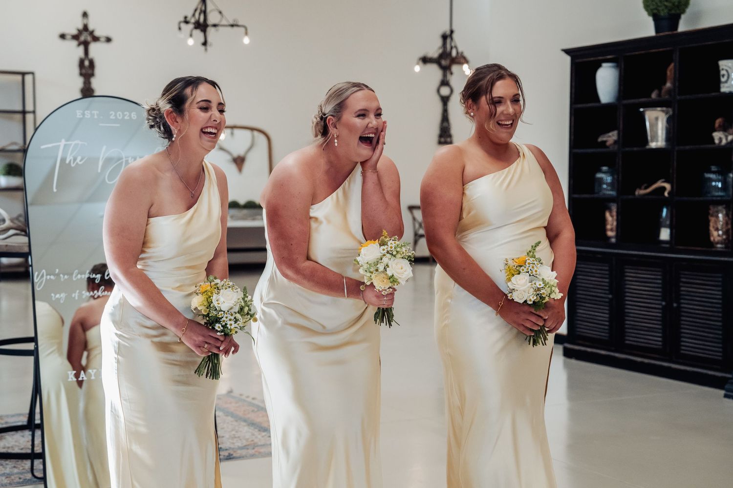 Three bridesmaids in ivory one-shoulder gowns laughing and holding white bouquets at a wedding ceremony.