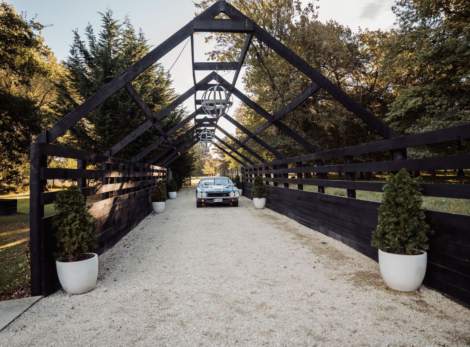 Car driving through elegant black wooden pergola entrance on gravel driveway.
