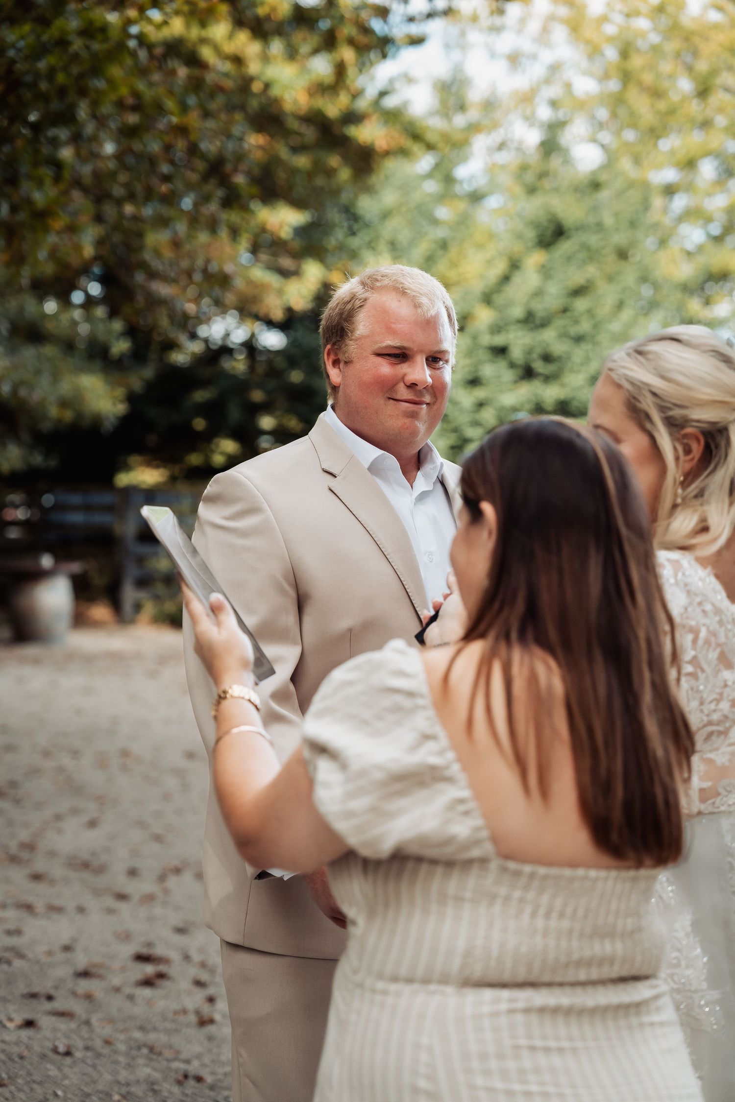 Groom in beige suit smiling during outdoor wedding ceremony while officiant reads vows.
