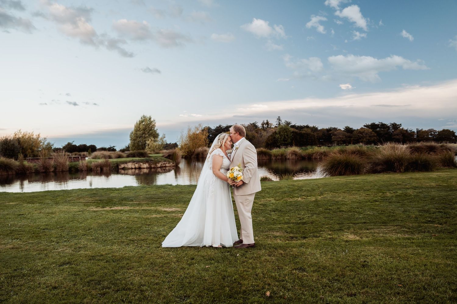 Bride and groom share a kiss near a serene pond surrounded by lush greenery under a beautiful blue sky.