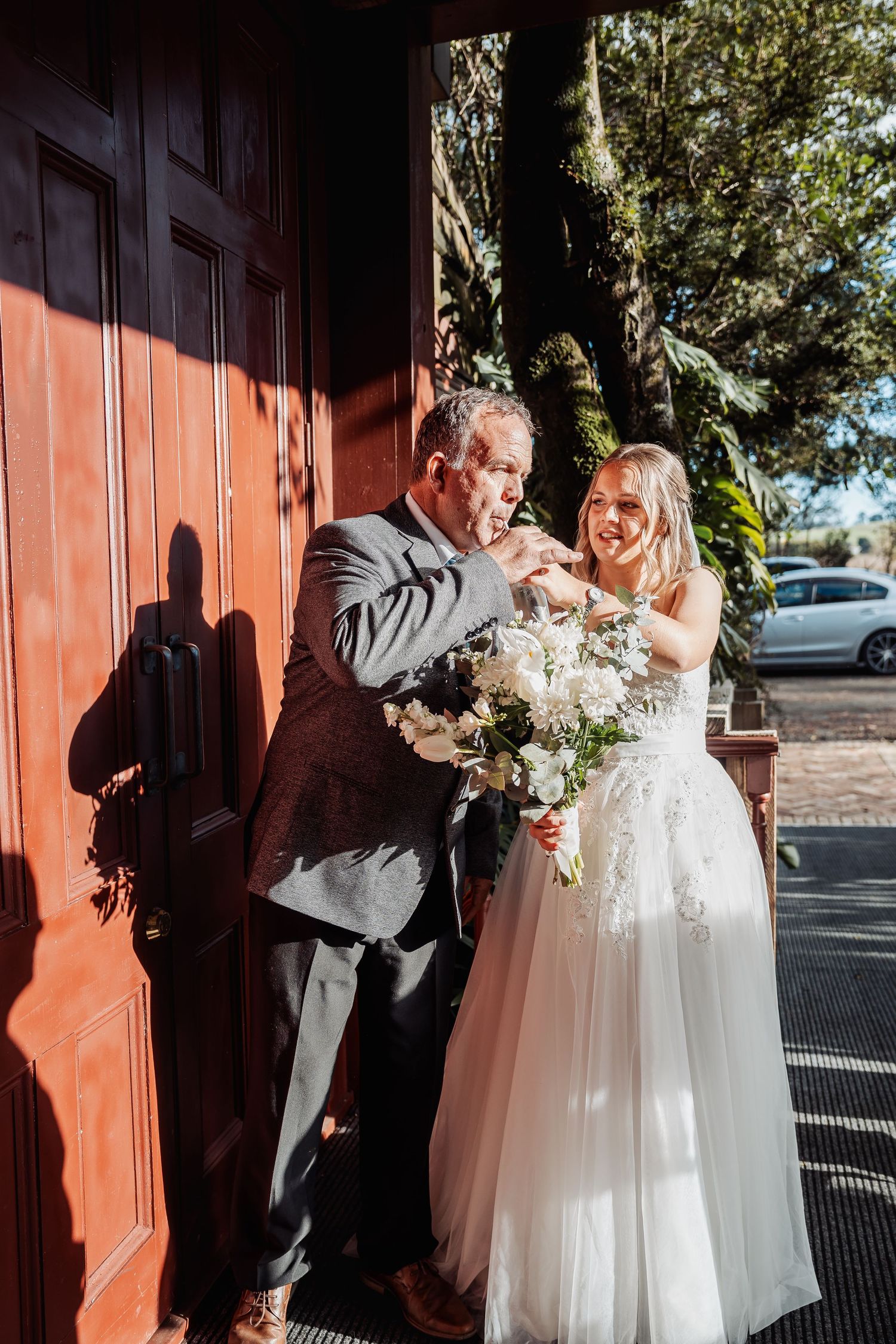 Wedding couple share joyful moment against red door while holding beautiful white floral bouquet.