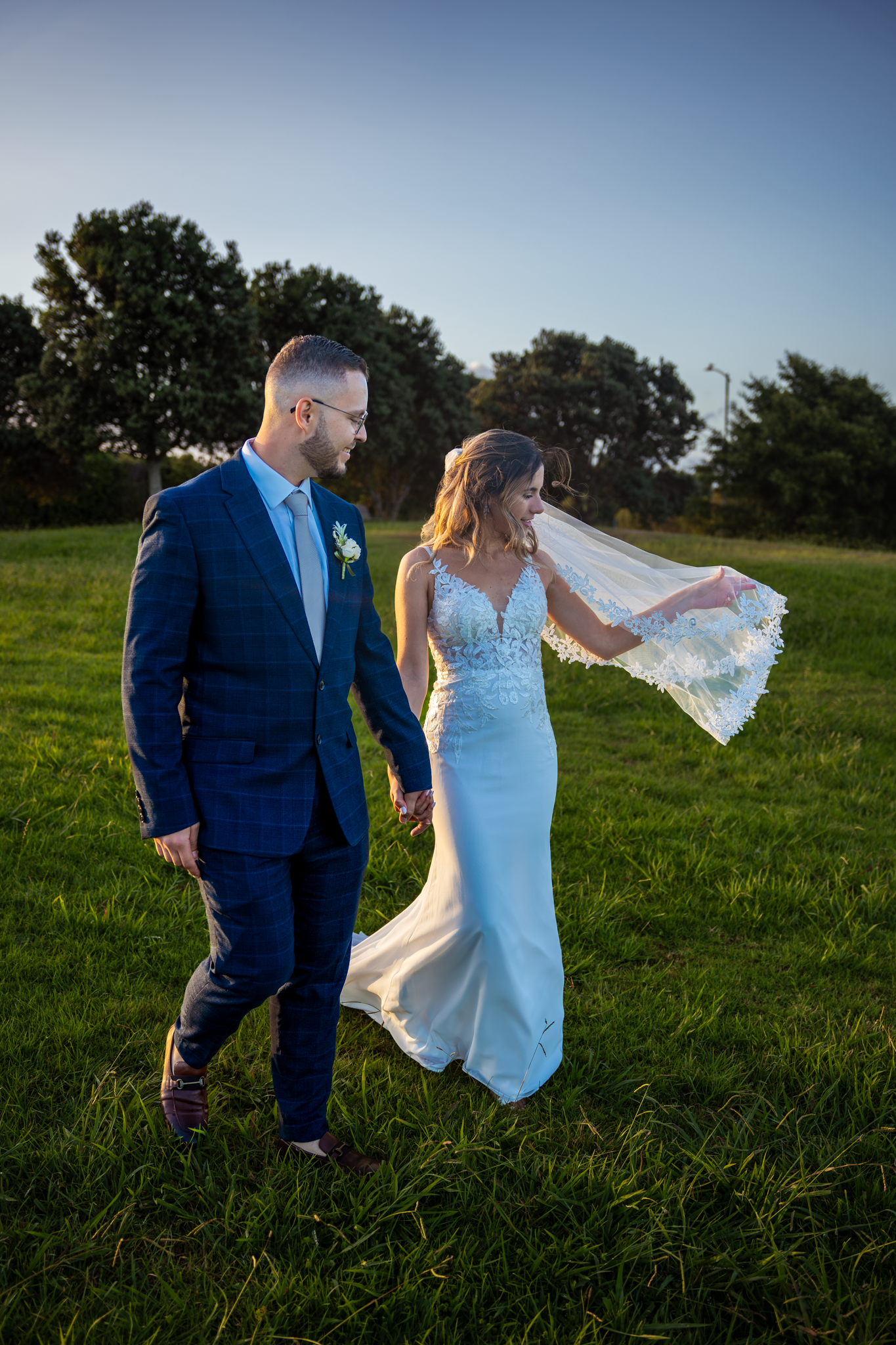 Bride and groom walking together through a grassy field at sunset.