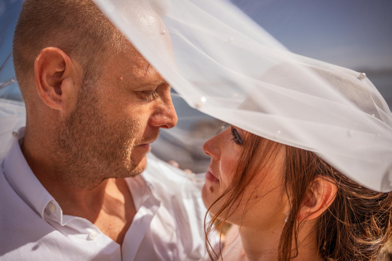 A close-up romantic moment captured under sunlight with white veil flowing in the breeze.