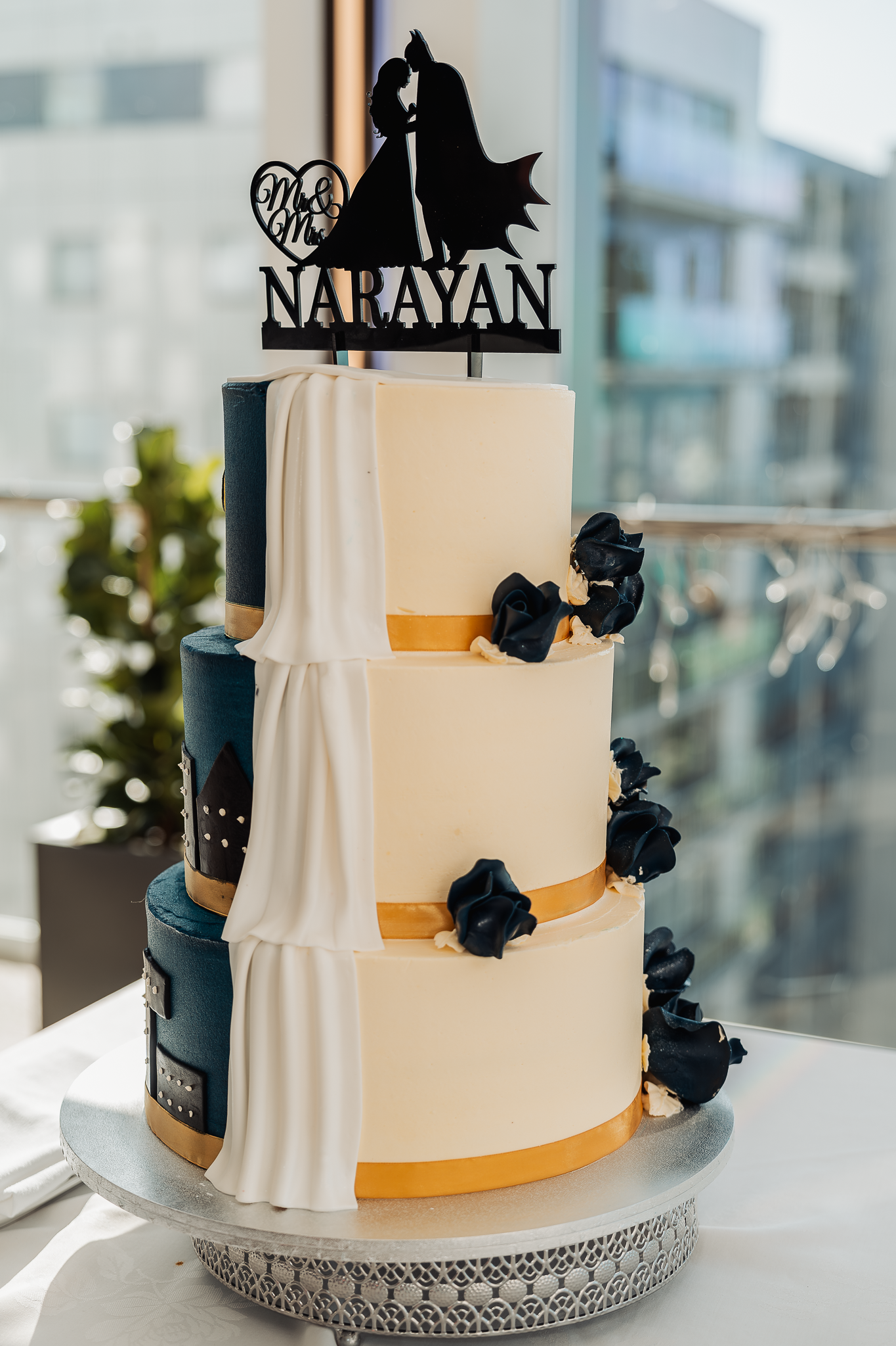 Three-tier white and navy wedding cake with gold bands and black flowers topped with a Batman silhouette cake topper.